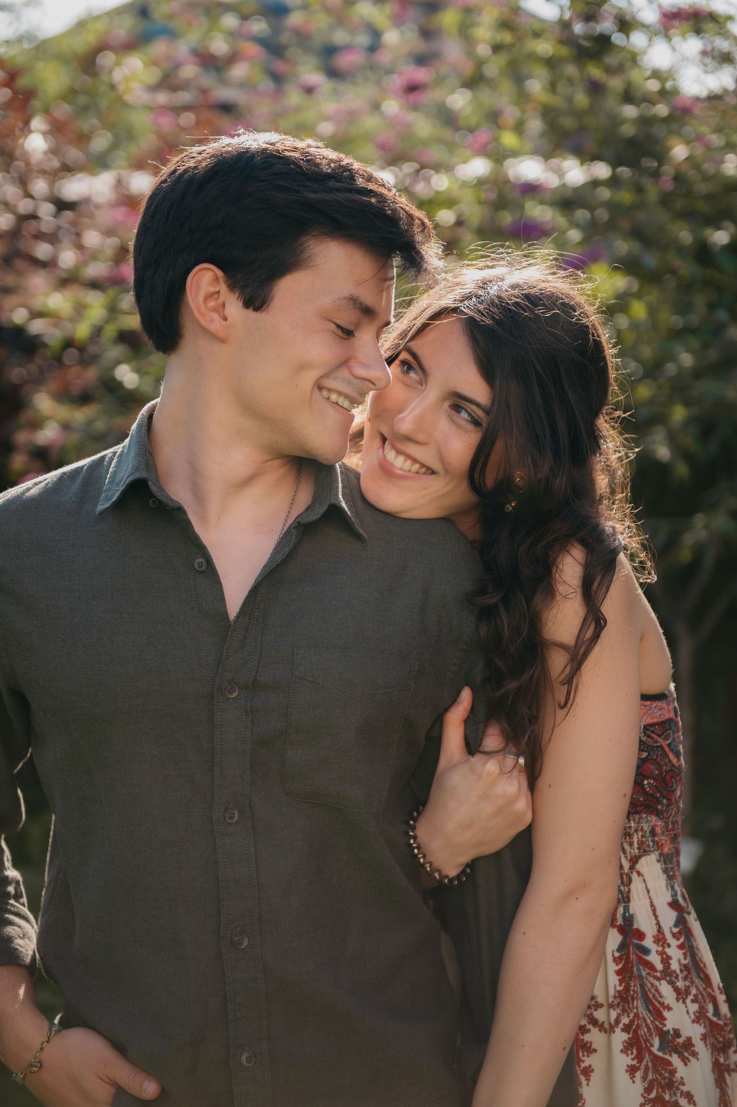 A smiling couple embraces outdoors with blooming flowers in the background, sharing a romantic moment in warm sunlight.