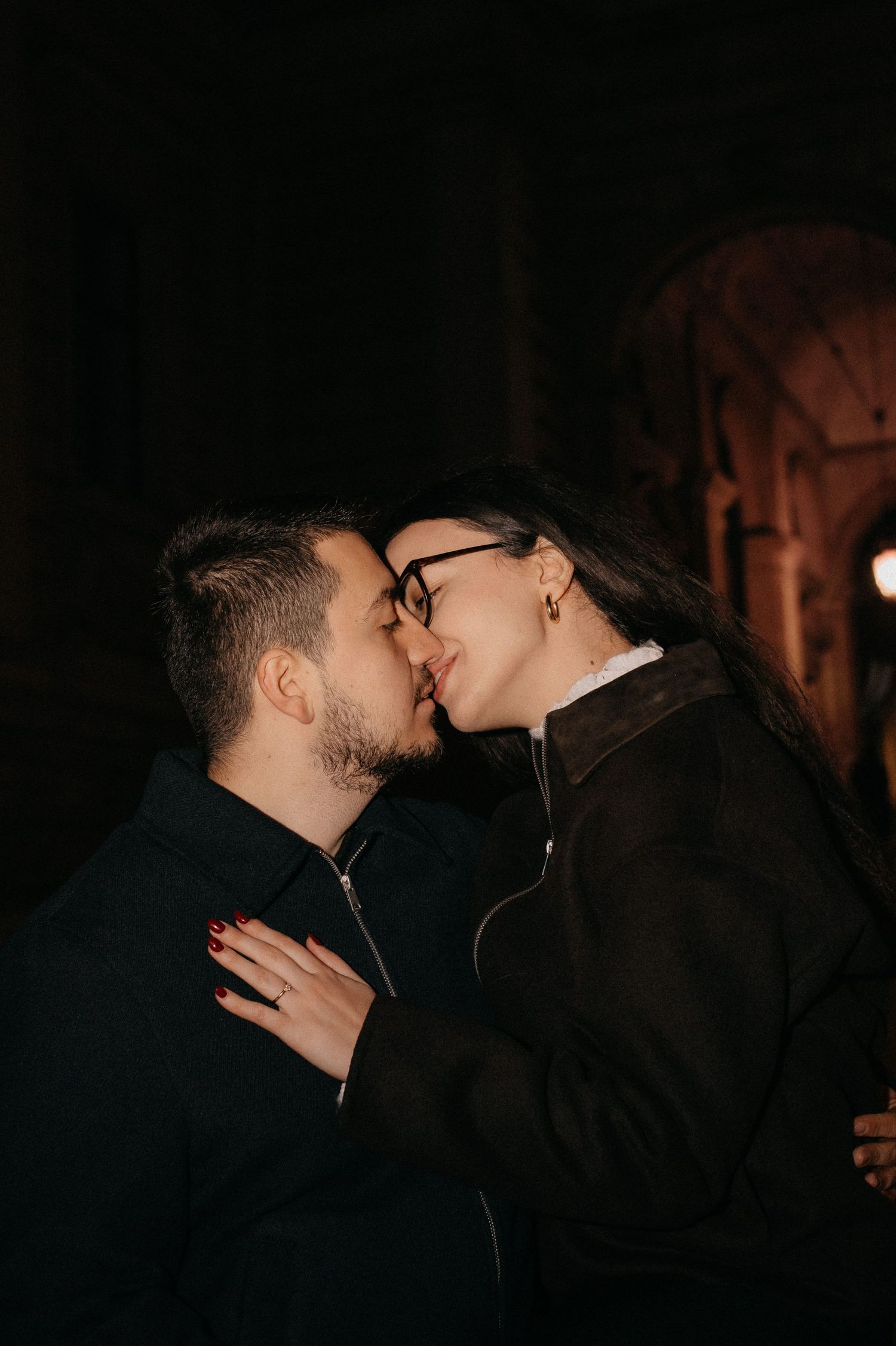 Couple sharing a romantic kiss at night against a dark stone archway backdrop during engagement session.