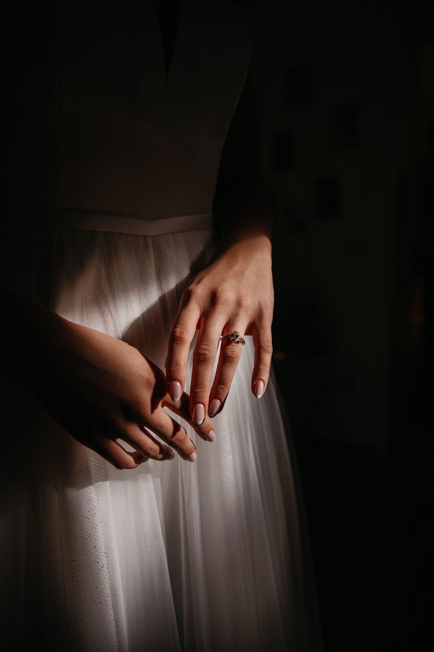 Close-up of bride's hands wearing a gold ring against her white tulle wedding gown in dramatic light.