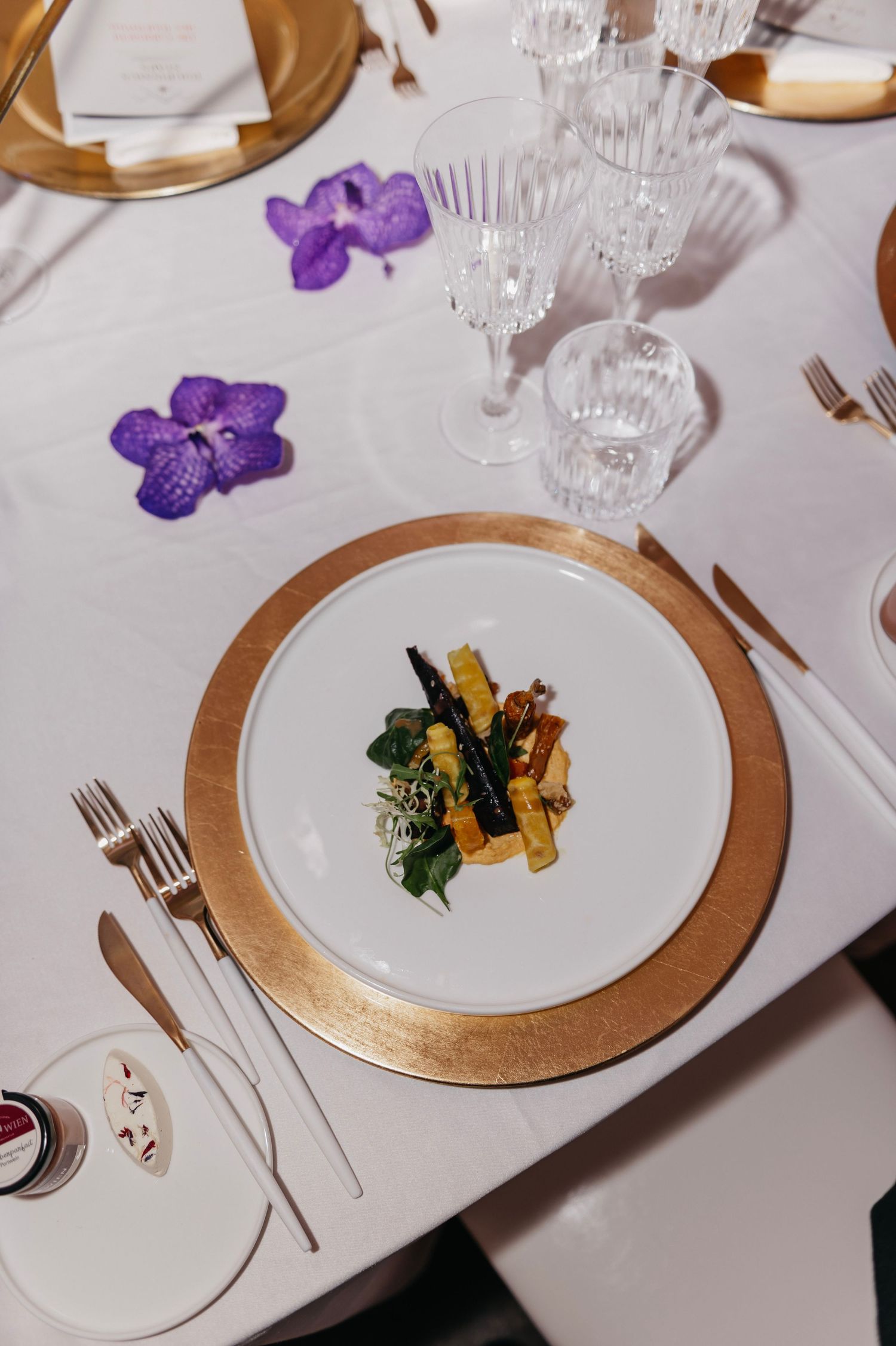 Elegant wedding place setting with gold charger plate, crystal glasses, purple orchids, and a gourmet appetizer on white linen.