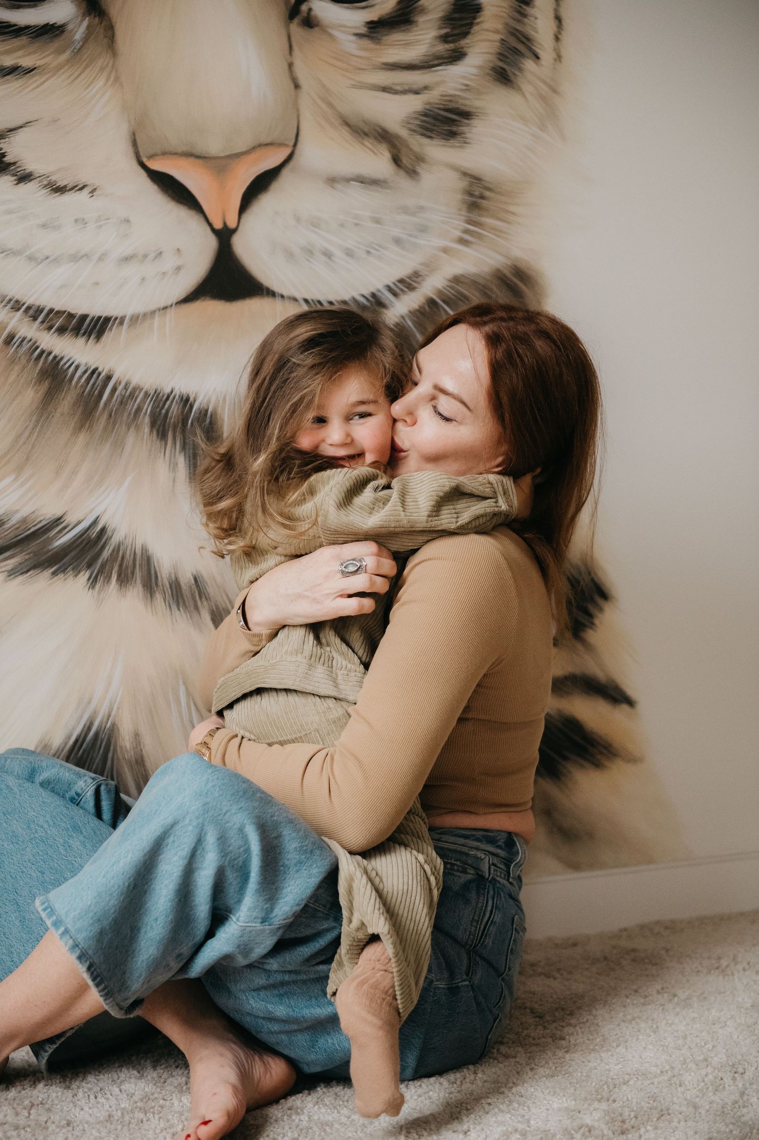 A mother and young daughter share a warm hug in front of a white tiger mural, sitting on a cozy carpet floor.