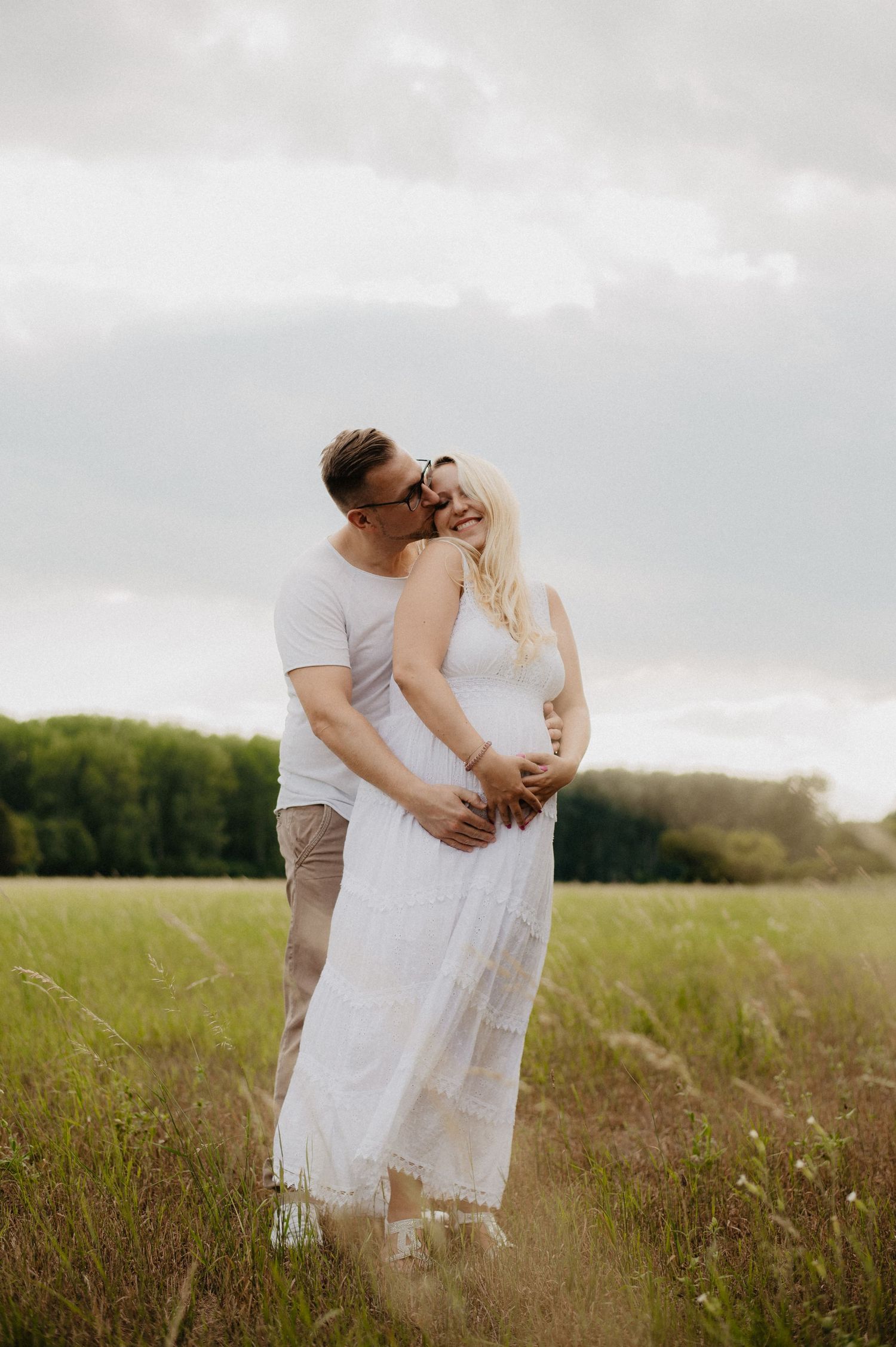 Couple shares tender kiss in green field, man embraces pregnant woman in white dress during outdoor maternity photo session.