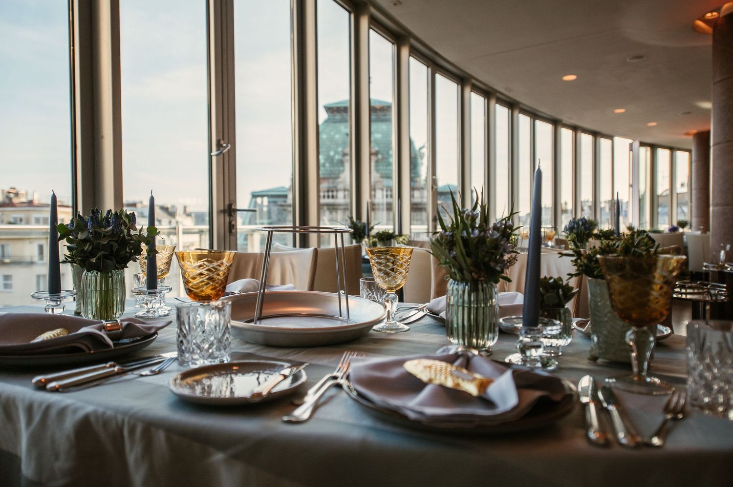 Elegant restaurant table setting with crystal glasses, floral centerpieces, and city views through curved floor-to-ceiling windows.