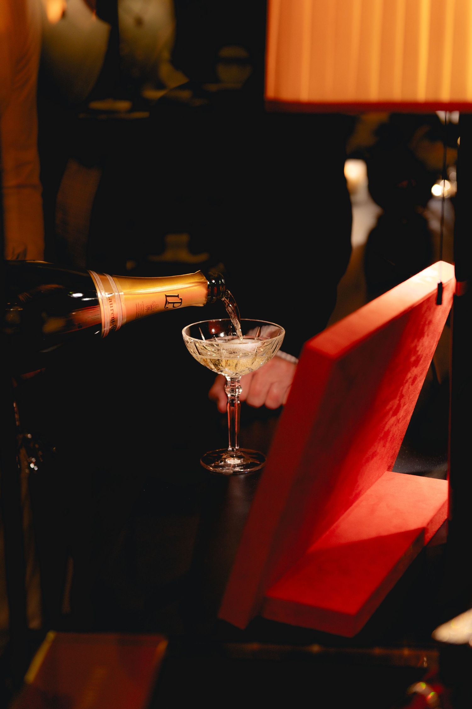 Champagne being poured into a coupe glass on a dark bar table beside a red velvet chair.