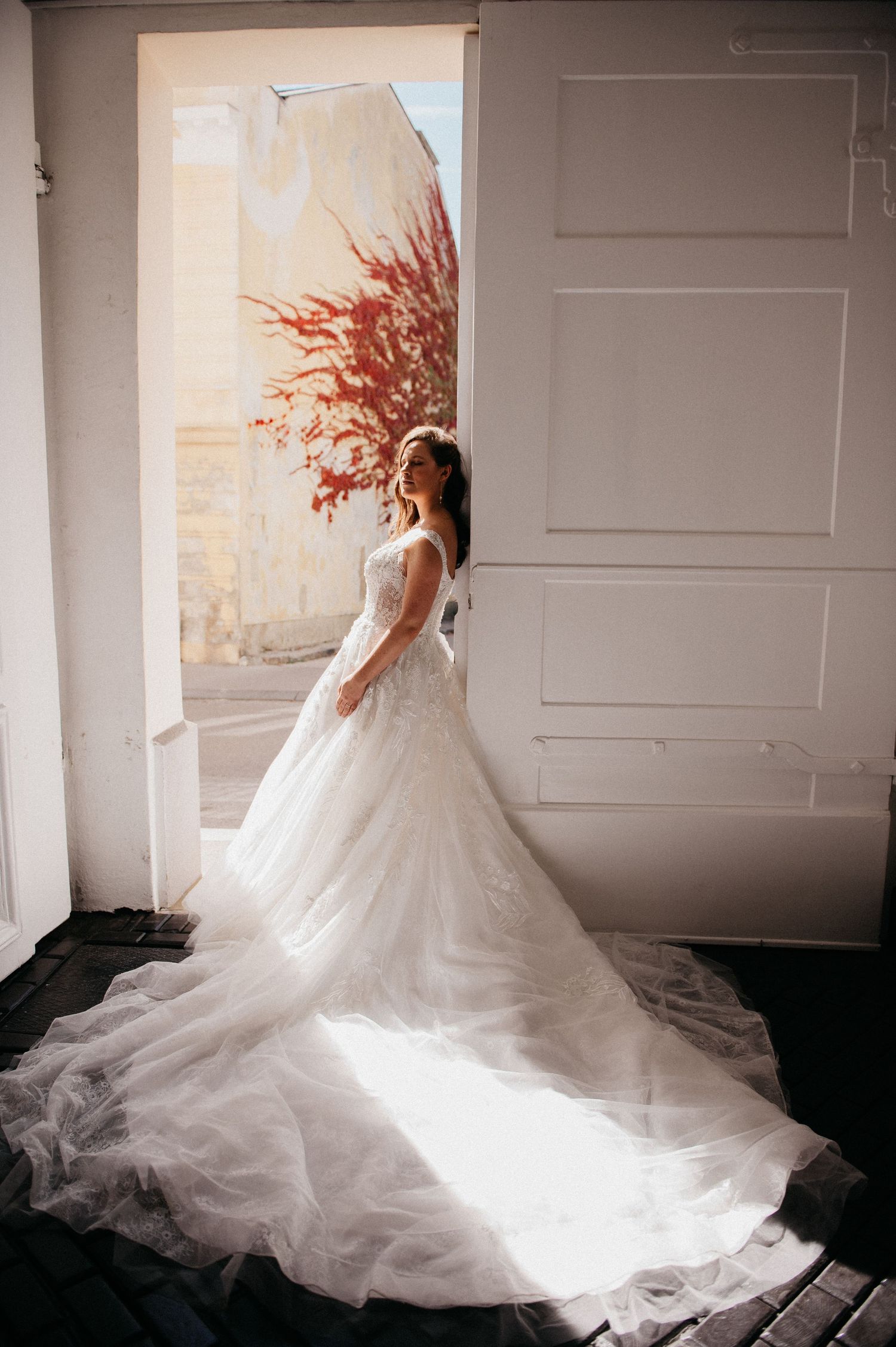 Bride in elegant white ball gown with long train stands in sunlit doorway with red tree backdrop.