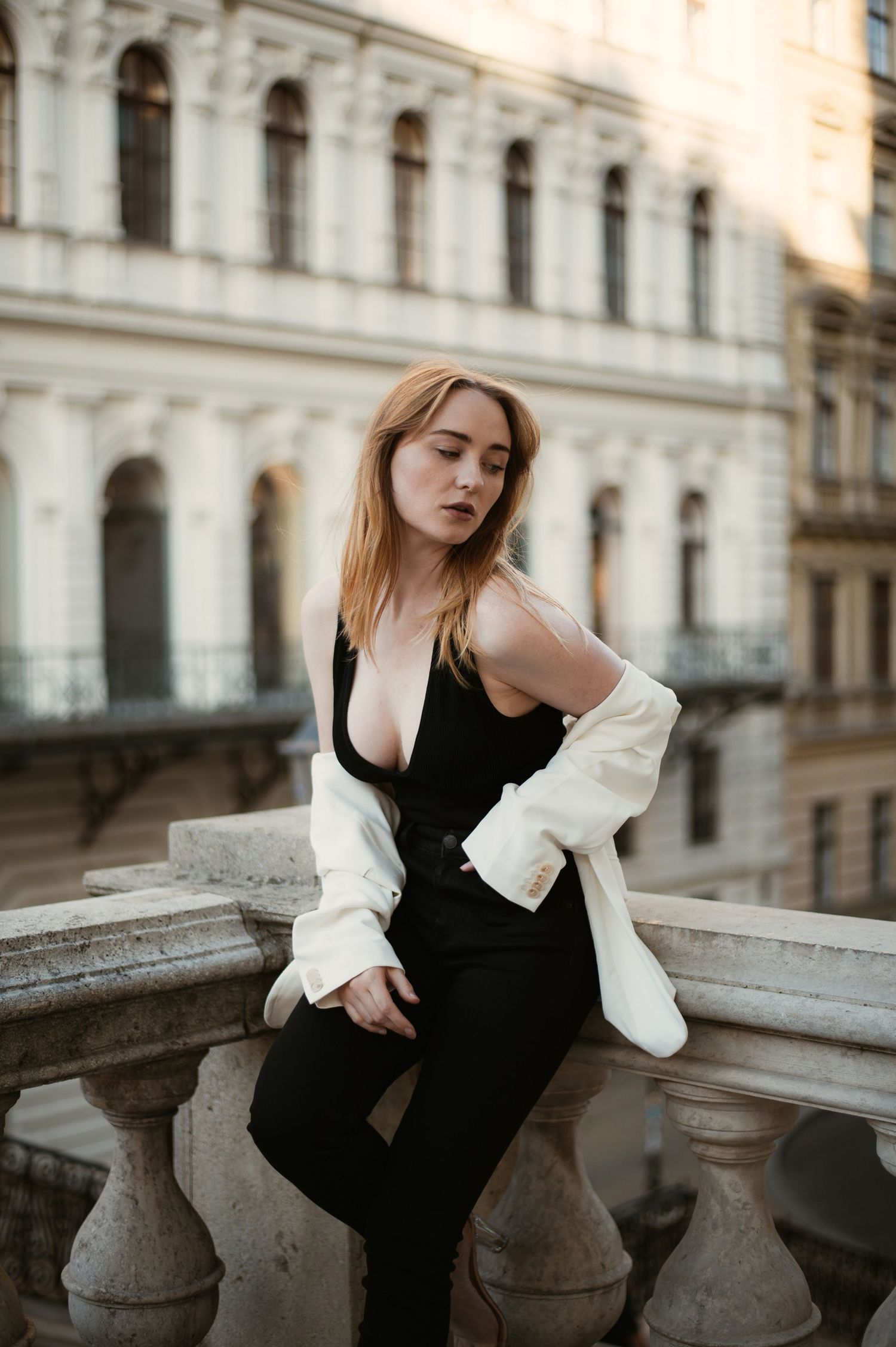 Blonde woman in black bodysuit and white jacket sits on ornate stone balcony railing of historic building.