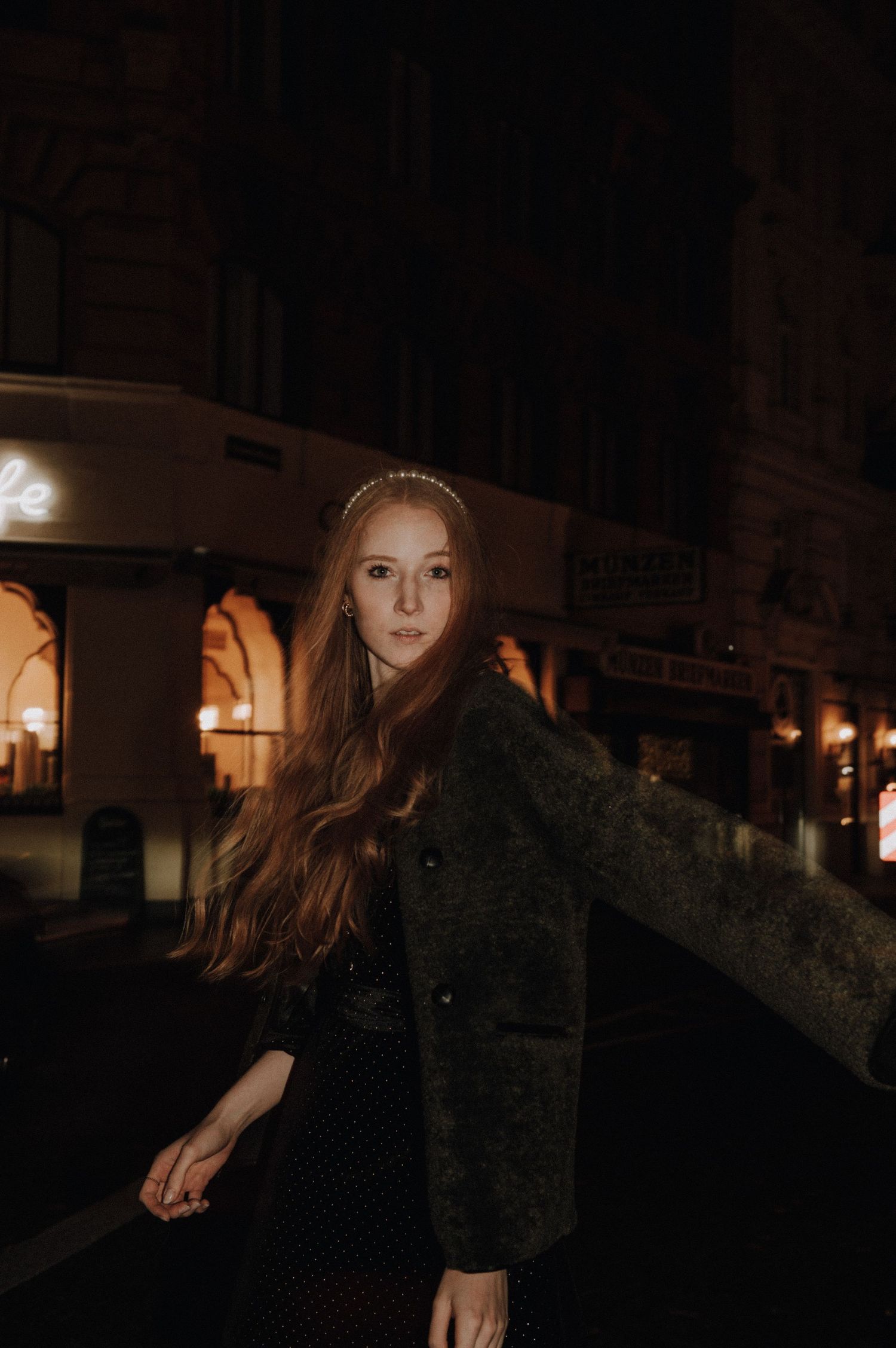 Young woman with long red hair wearing a dark green coat poses on a city street at night.