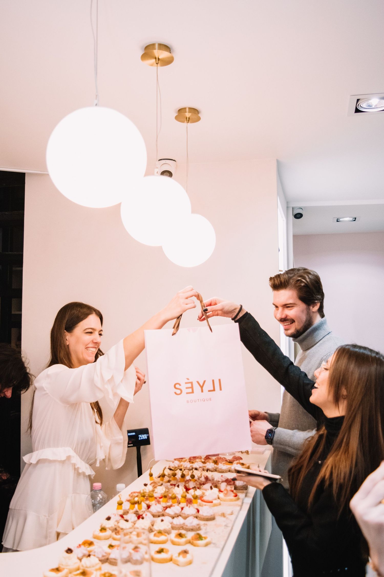 Smiling guests exchange a pink ILYES boutique shopping bag at an elegant indoor event with pastries.