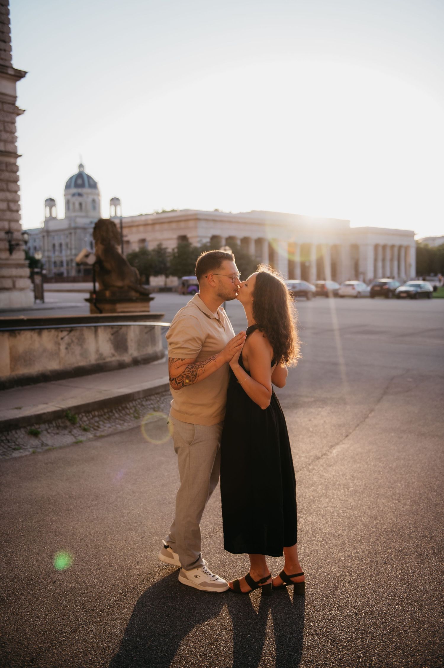 Couple sharing a kiss at golden hour in front of a grand European museum with sunlight flare.