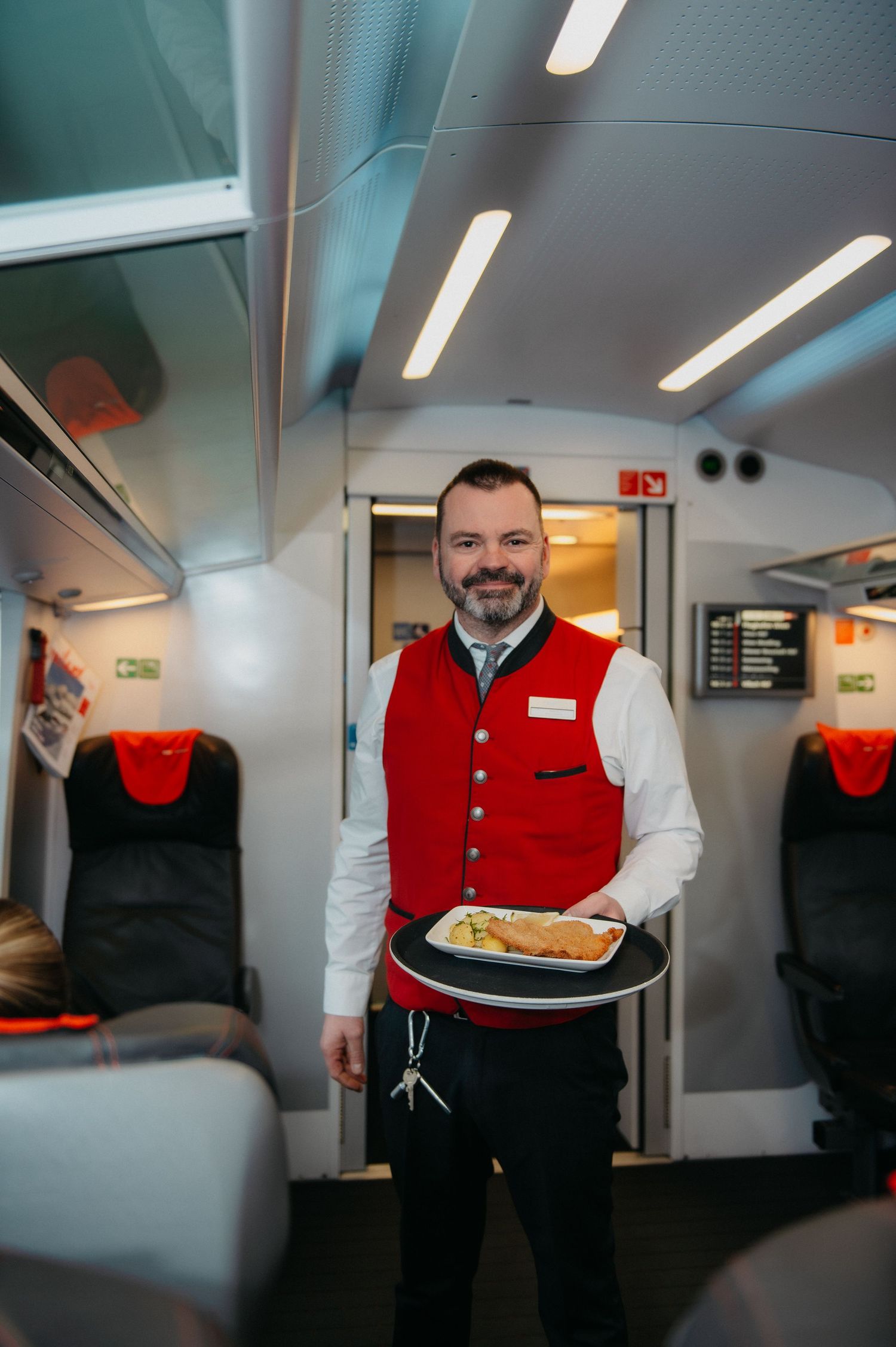 A smiling train attendant in a red vest carries a meal tray through a modern passenger train car aisle.