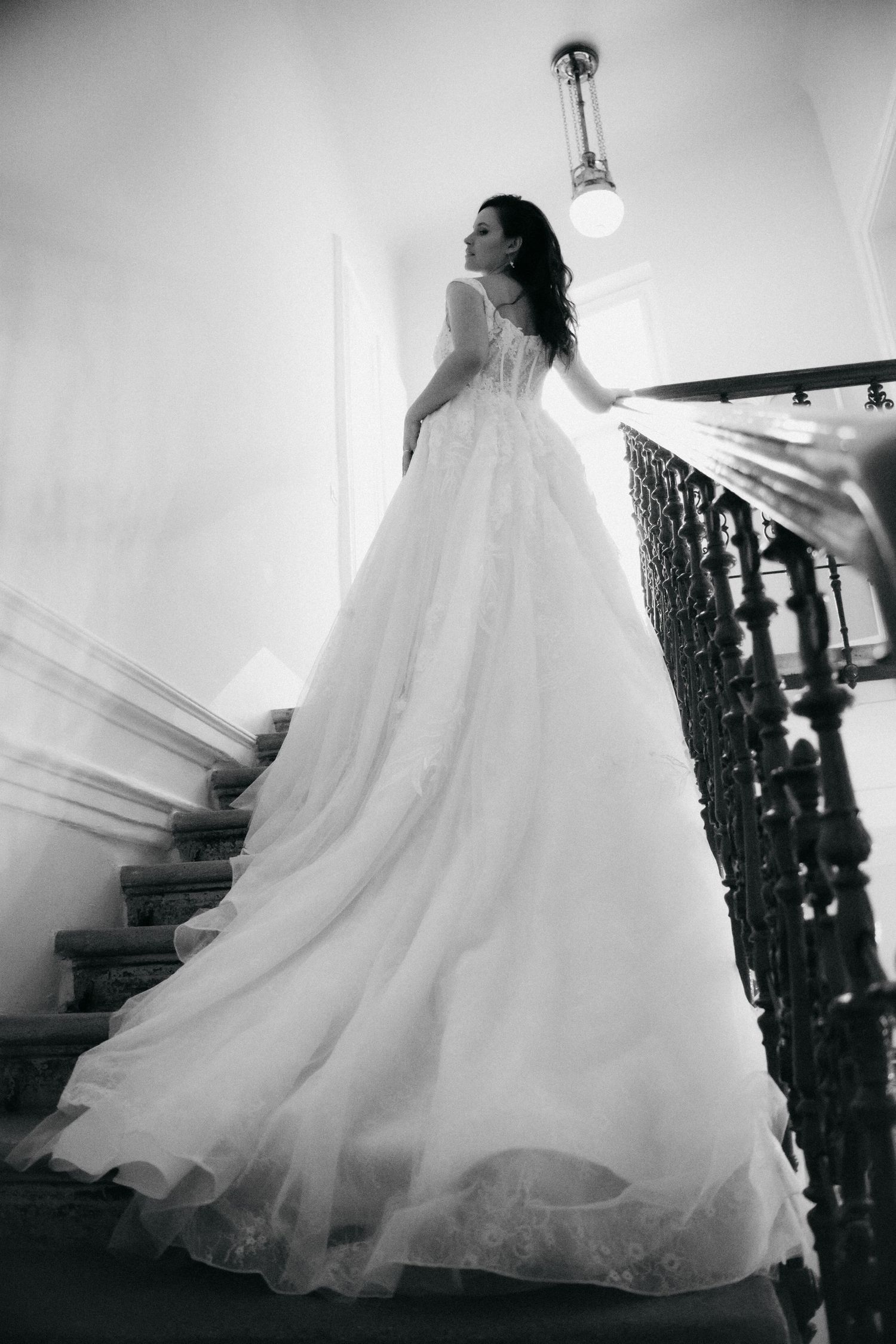 Elegant bride in a flowing white ball gown stands on ornate staircase, black and white wedding photography.