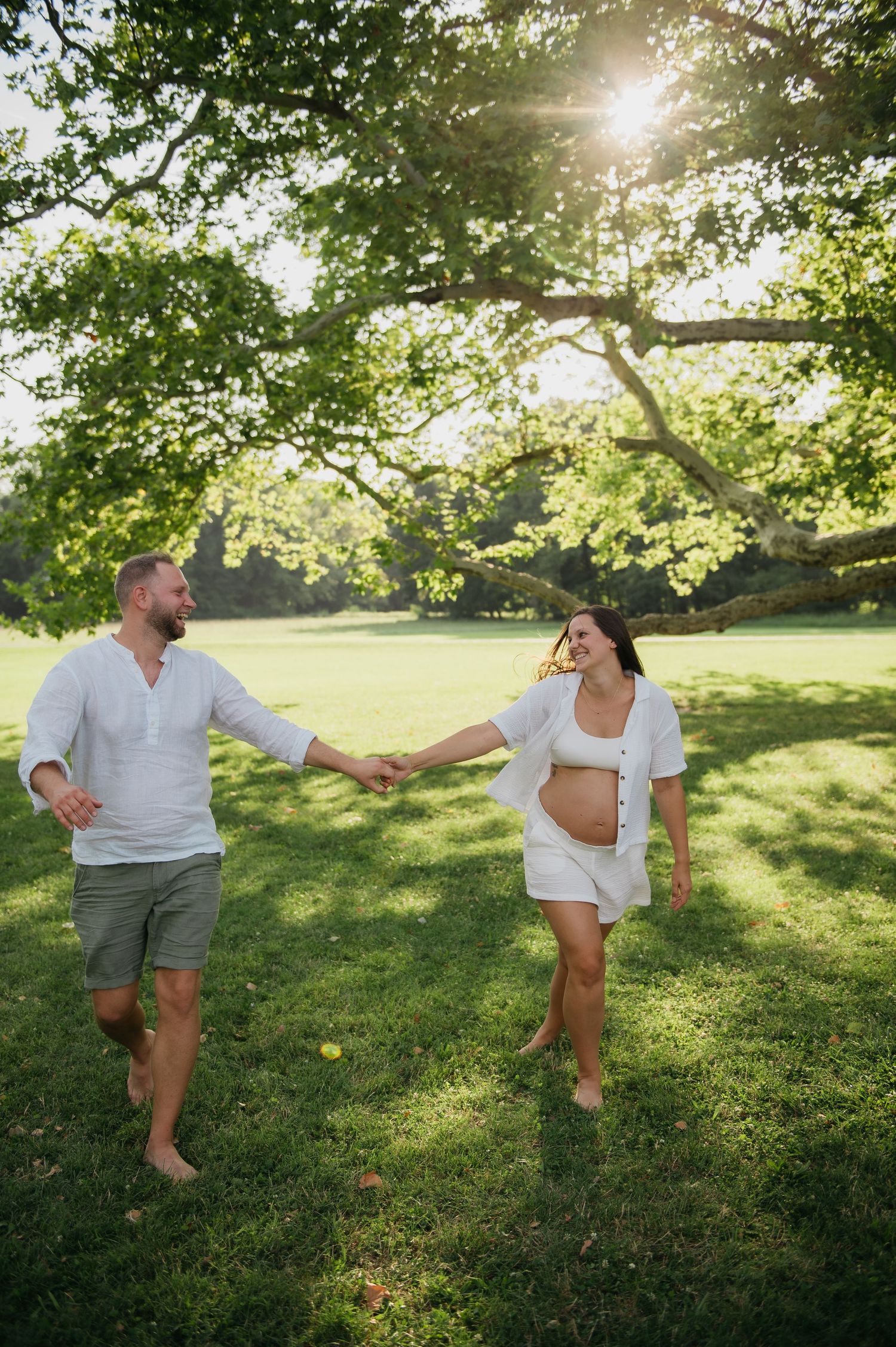 Pregnant couple holding hands and walking barefoot on green grass under sunlit trees during an outdoor maternity photoshoot.