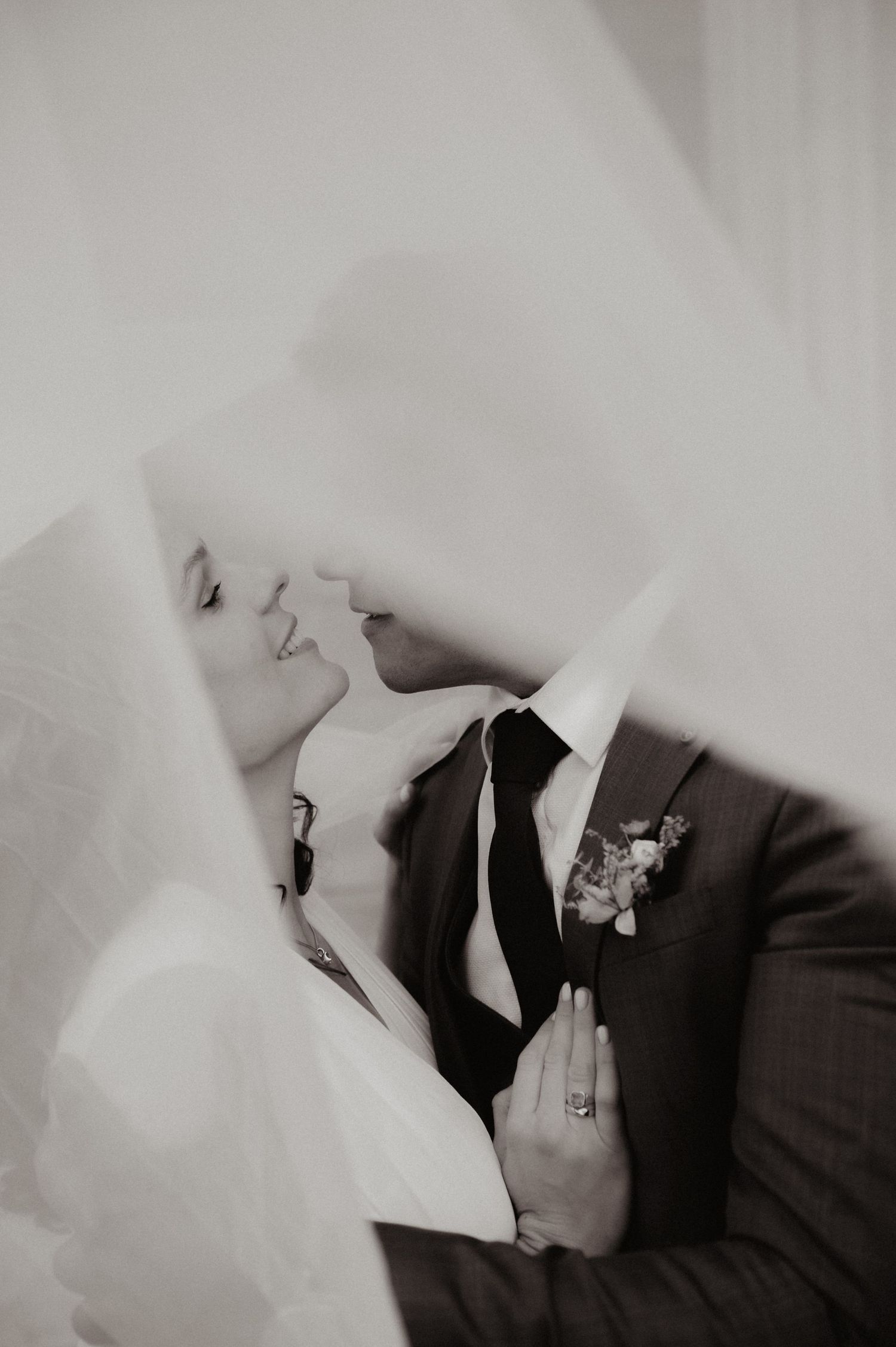 Black and white photo of bride and groom sharing a romantic kiss under a flowing veil on their wedding day.