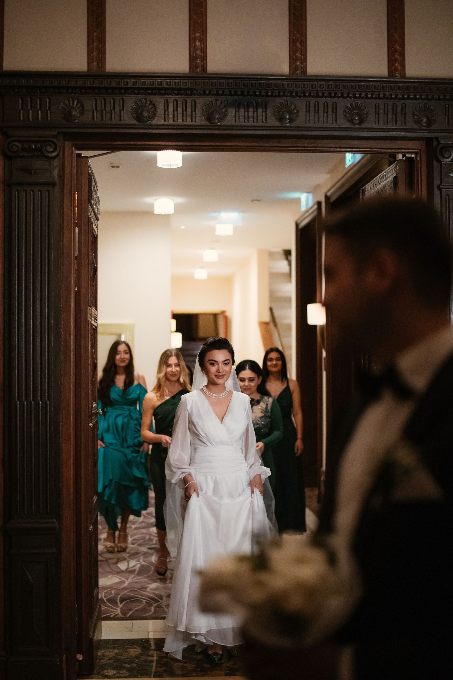 Bride in white dress walks through ornate doorway followed by bridesmaids in green dresses, groom visible in foreground.