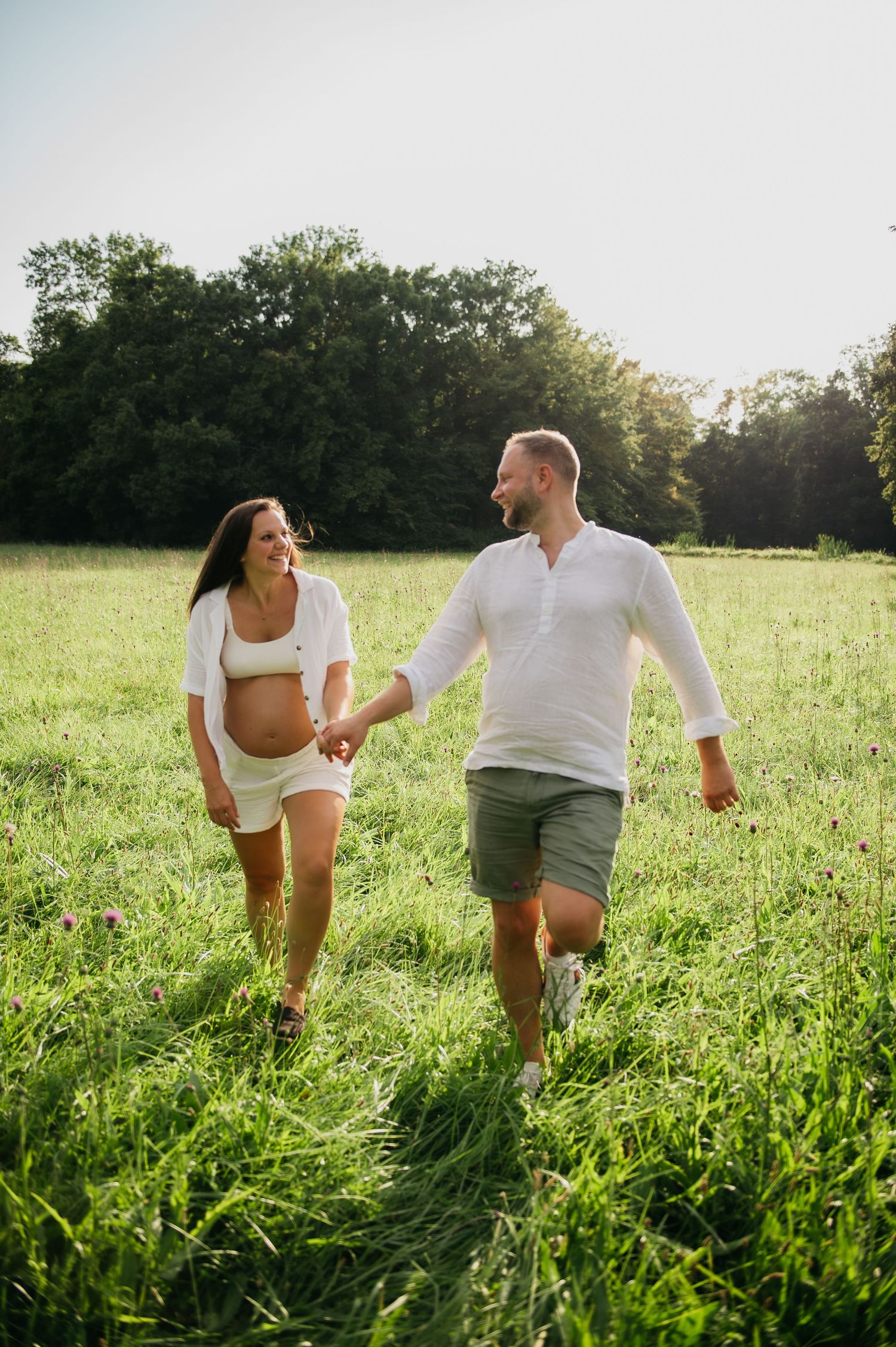 Pregnant couple holding hands and walking through a sunny green meadow, smiling at each other in matching white outfits.
