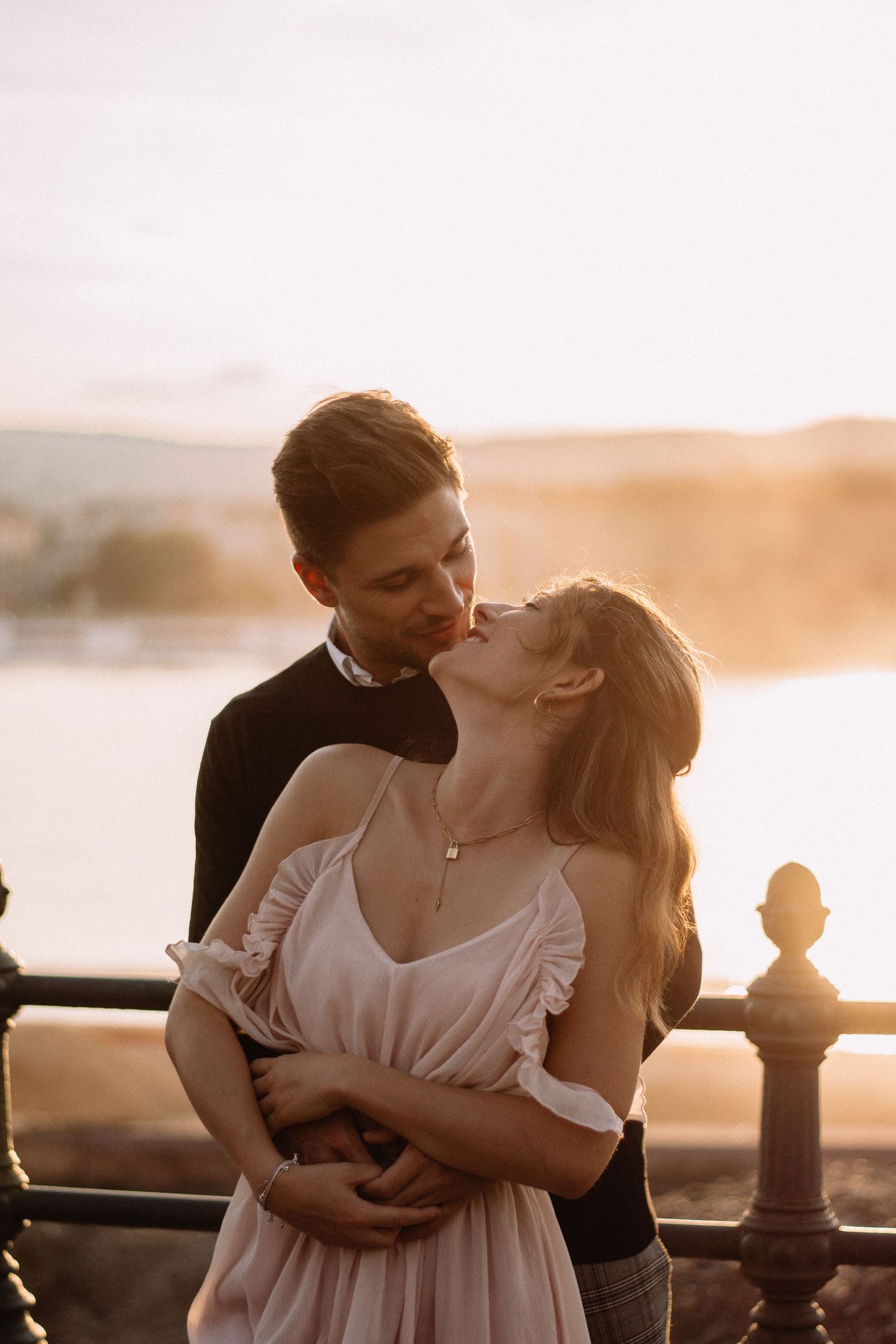 Couple sharing a romantic kiss at sunset by a waterfront railing, woman in white dress embraced by man in dark outfit.