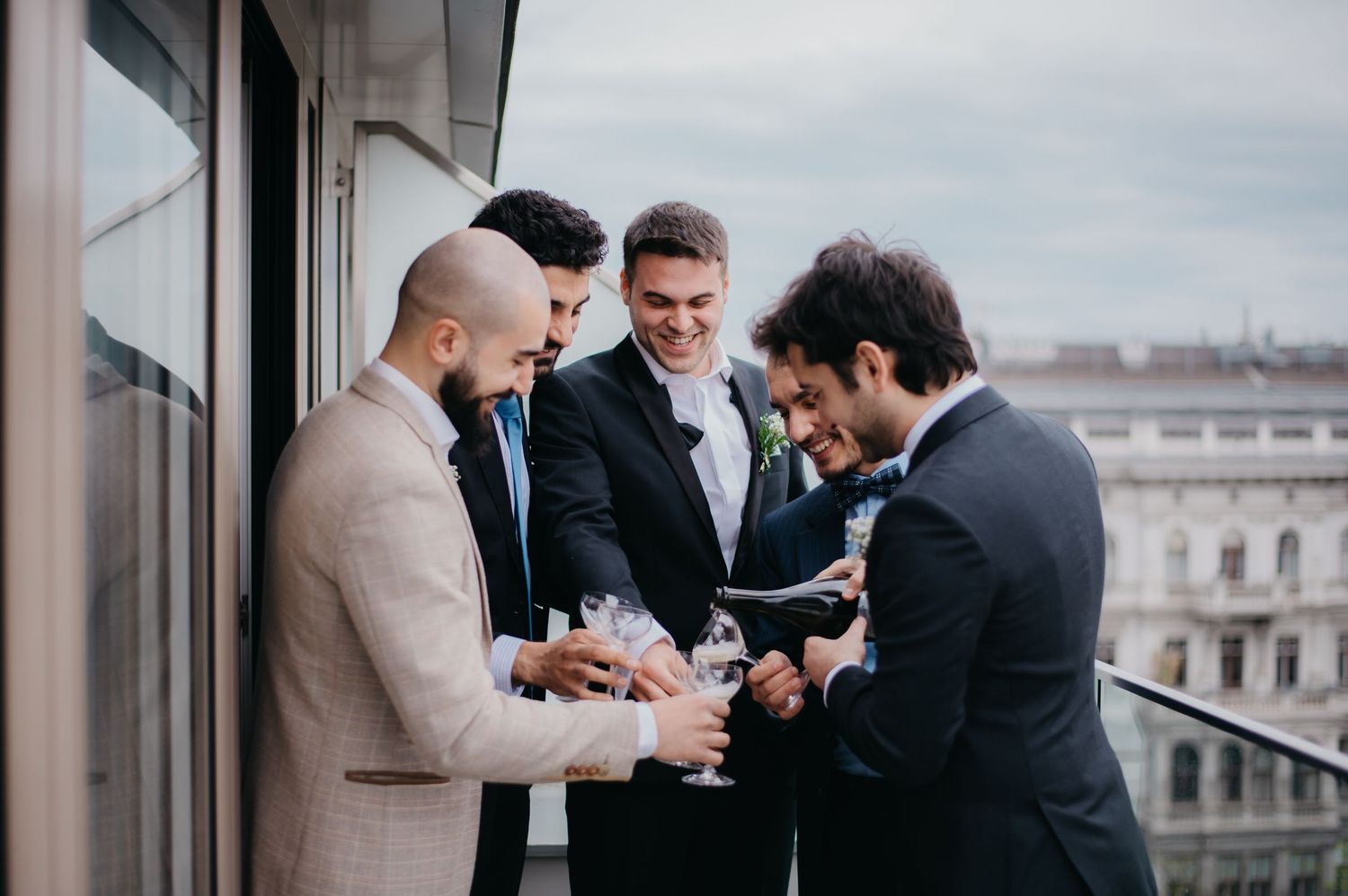 Group of well-dressed men in suits celebrating on a balcony, toasting with champagne glasses at a formal event.