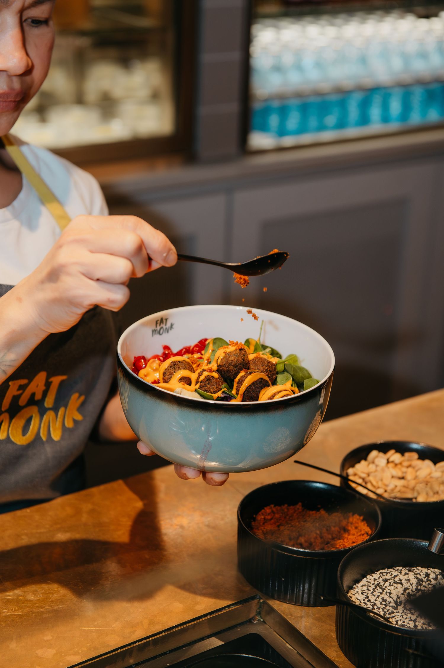 Chef drizzling sauce over a colorful ramen bowl at Fat Monk restaurant, with toppings of falafel, peppers, and greens.