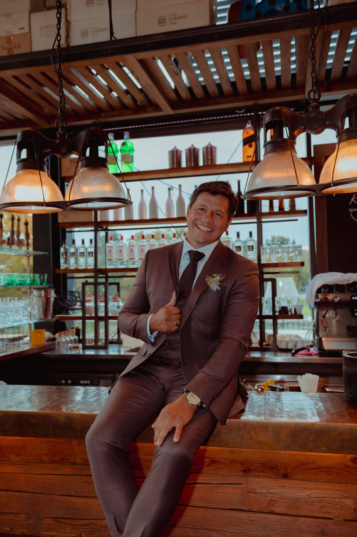 Smiling groom in brown suit with boutonniere sitting at a bar with pendant lights and liquor bottles behind him.