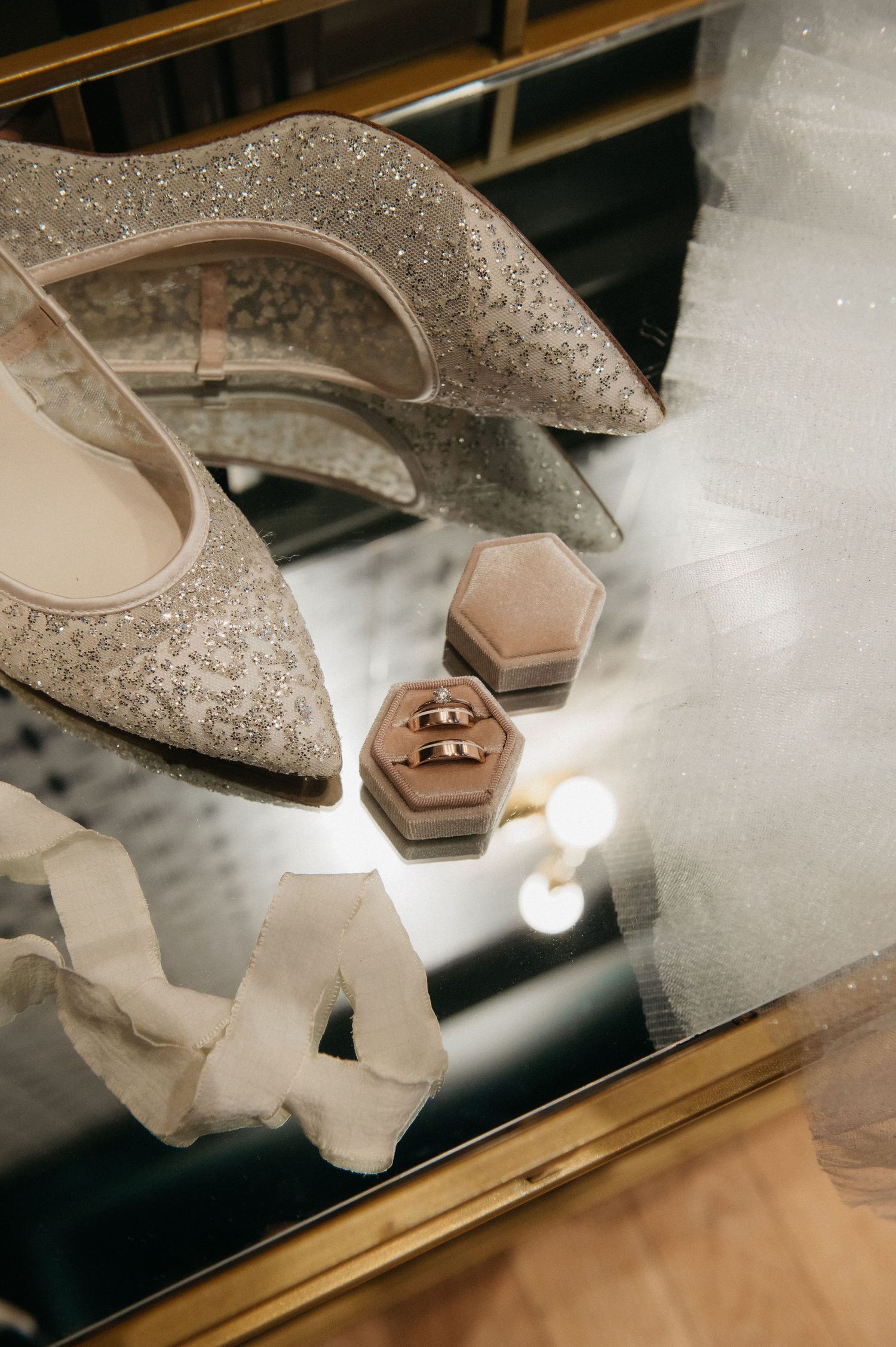 Glittering bridal heels, wedding rings in velvet box, and white veil arranged on mirrored surface.