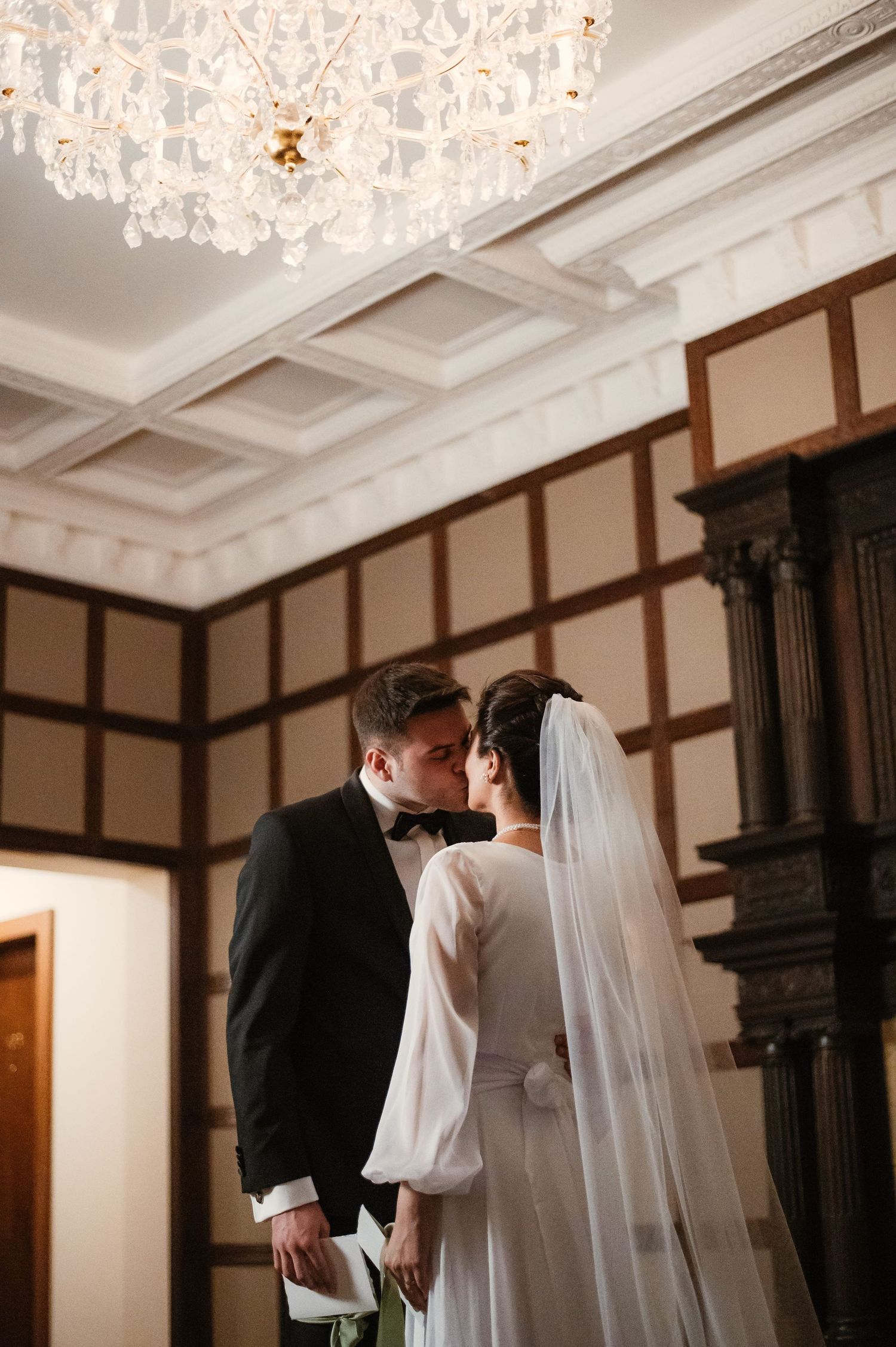 Bride and groom kissing beneath crystal chandelier in ornate wood-paneled wedding venue.