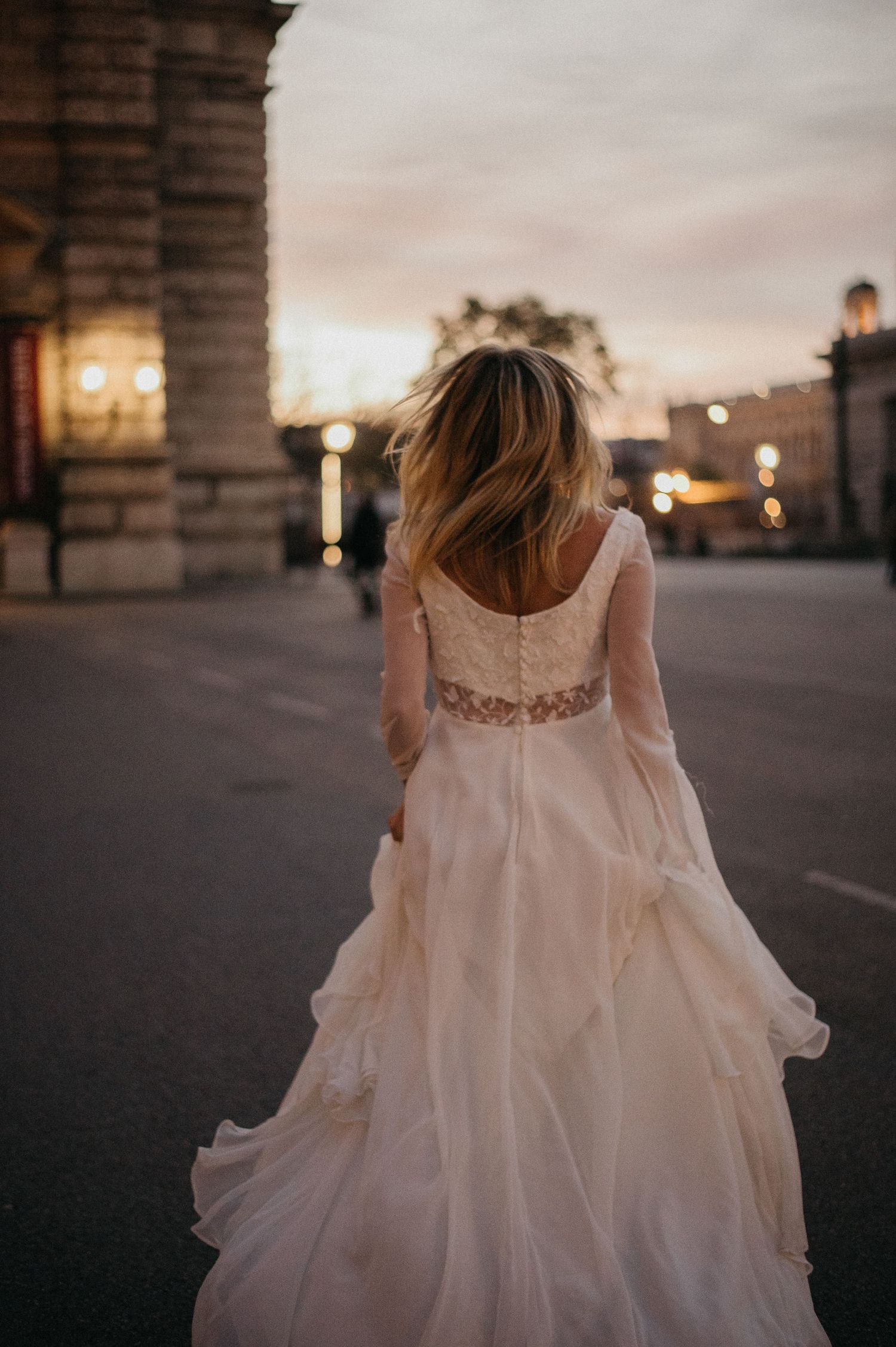 Bride in a flowing white lace wedding gown walks at sunset in an urban setting, hair blowing in the breeze.