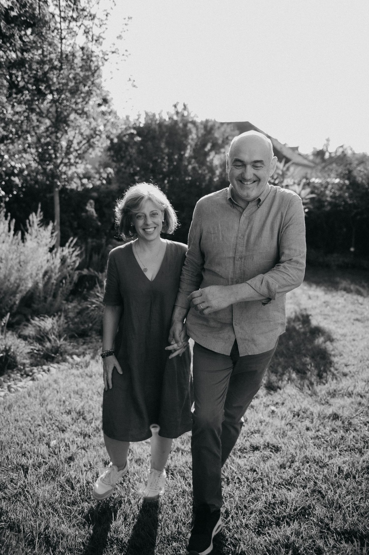 Black and white photo of a smiling couple standing together in a lush garden, casually dressed and enjoying the outdoors.