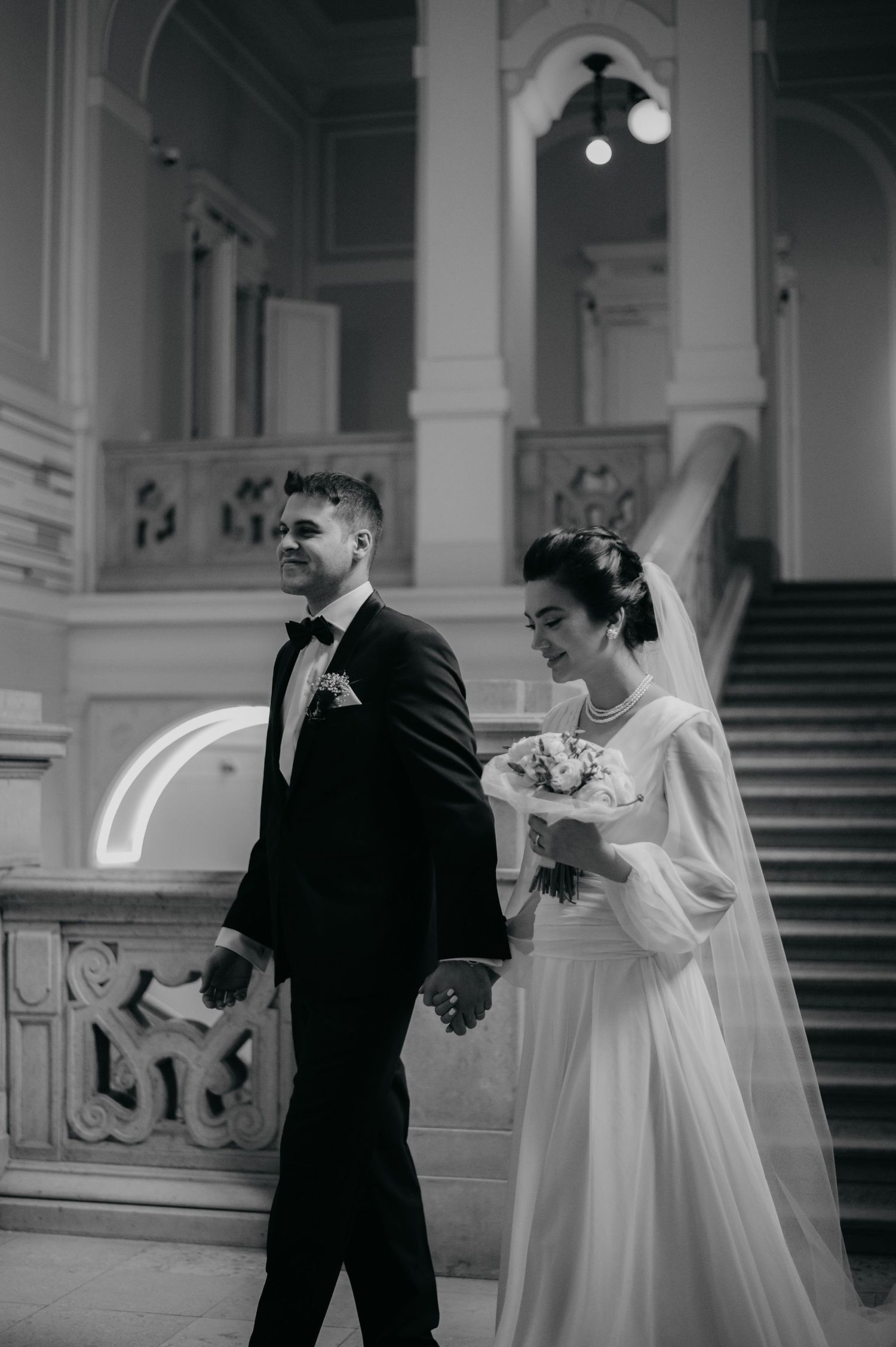 Black and white wedding photo of bride and groom holding hands walking through elegant grand hall with staircase.