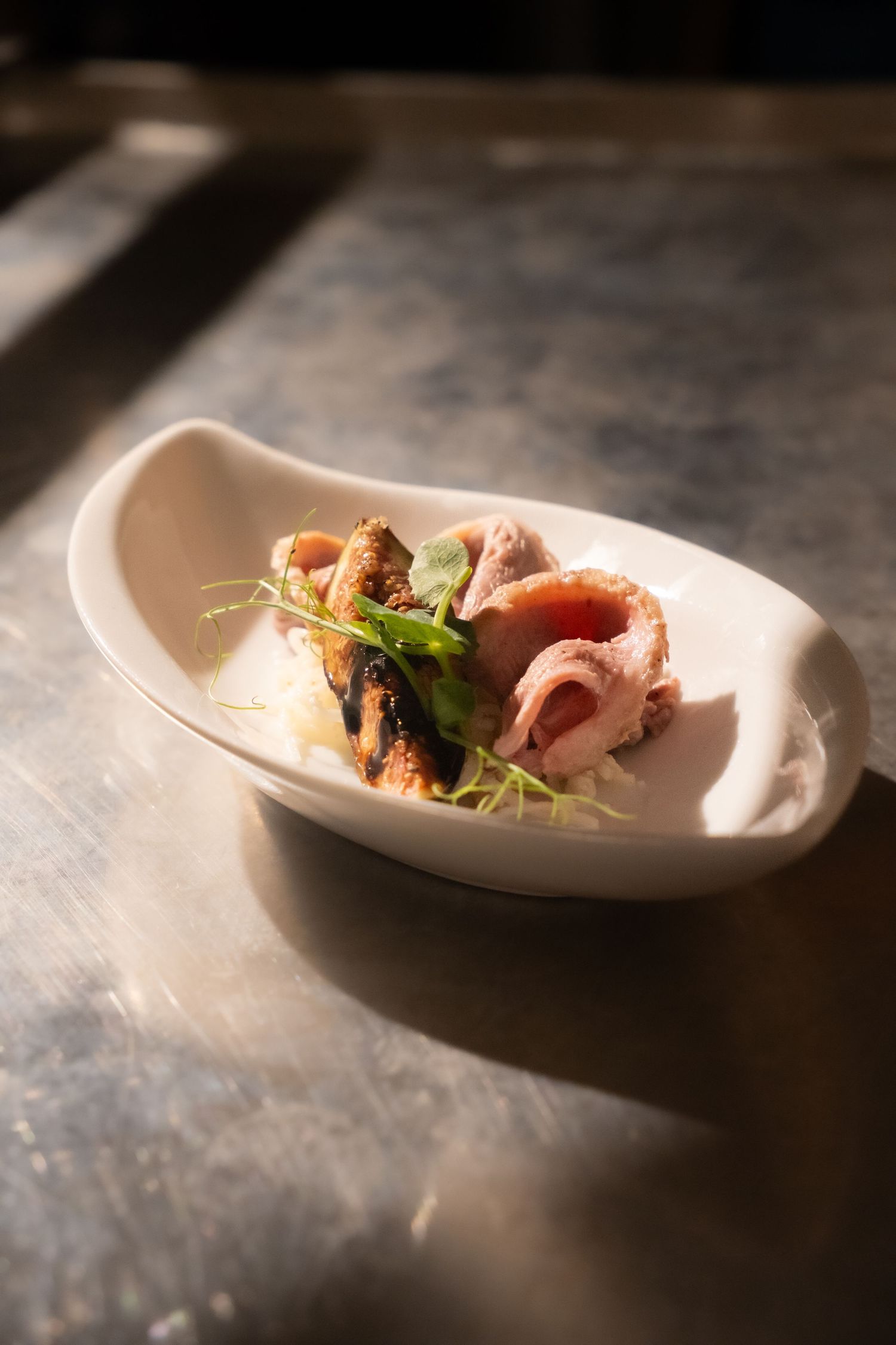 Elegant plated dish with sliced meat and fresh herbs served in a white oval bowl on a metal surface.