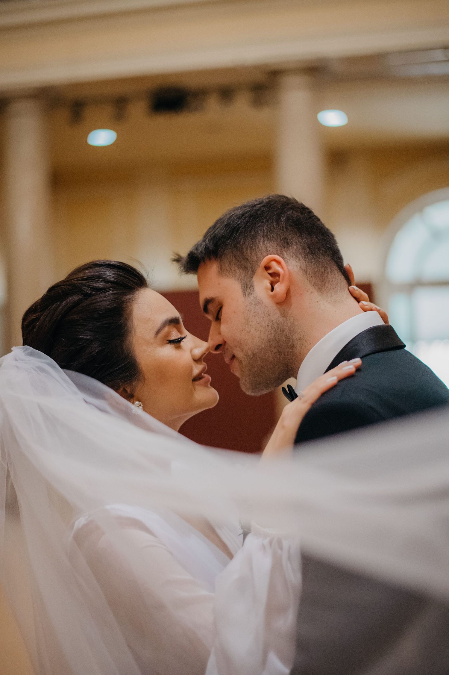 Bride and groom sharing intimate almost-kiss moment under flowing veil in sunlit church.