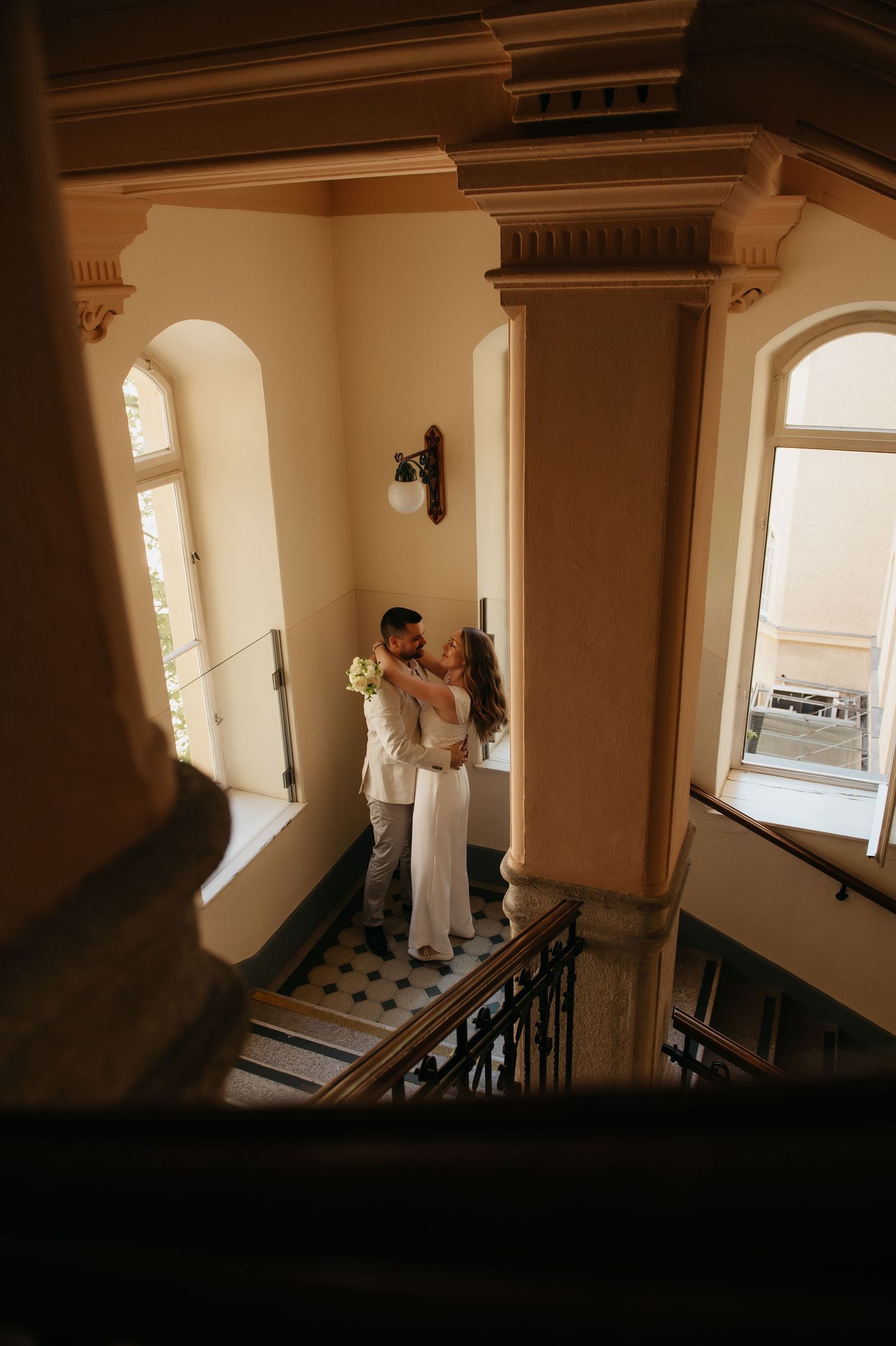 Couple embracing on elegant staircase landing in historic building, bride in white dress holding bouquet, groom in light suit.