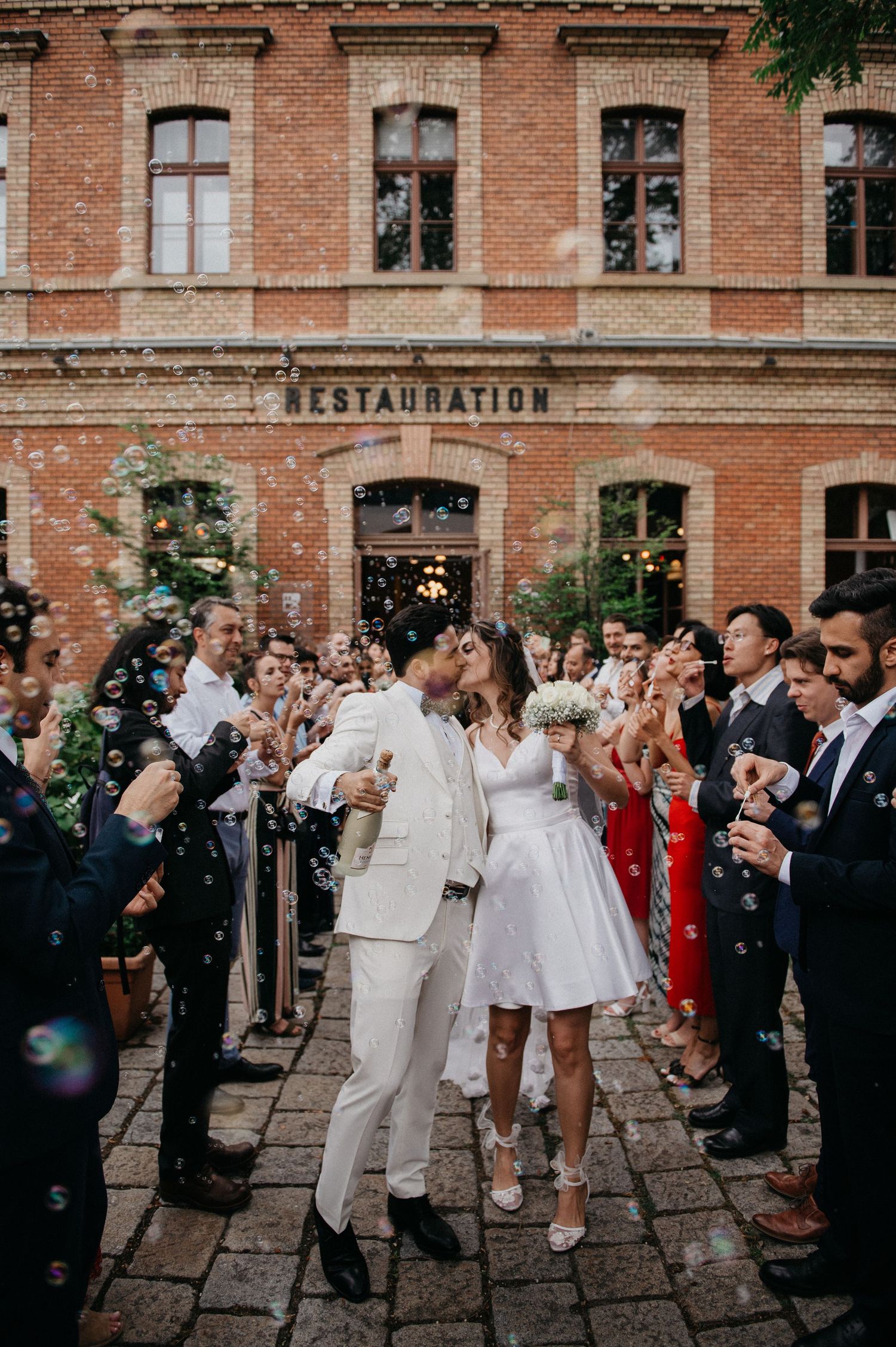 Bride and groom walk through bubble shower outside brick Restauration building surrounded by cheering wedding guests.