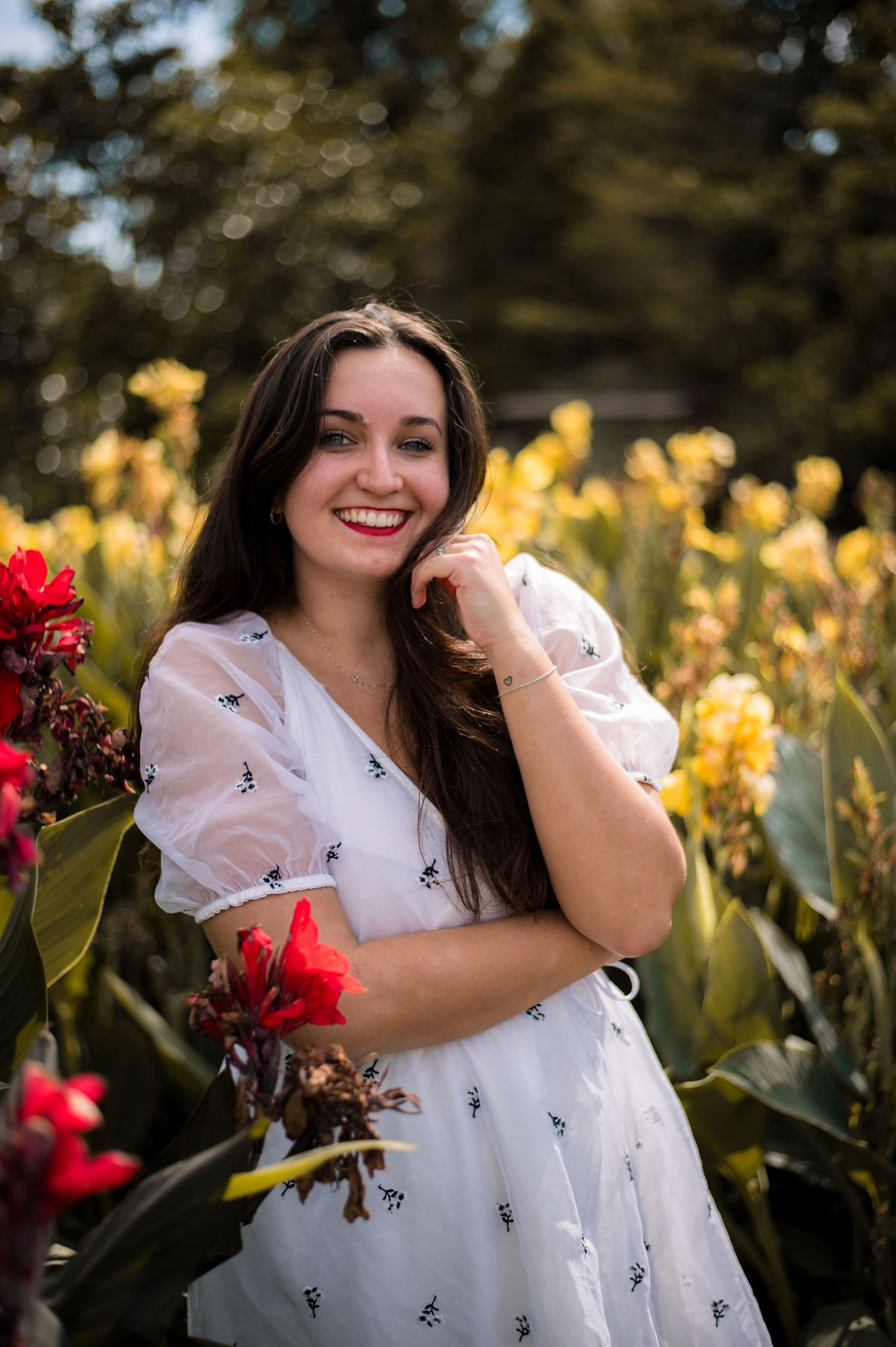 Young woman in white dress smiling among colorful red and yellow flowers in a sunny garden.