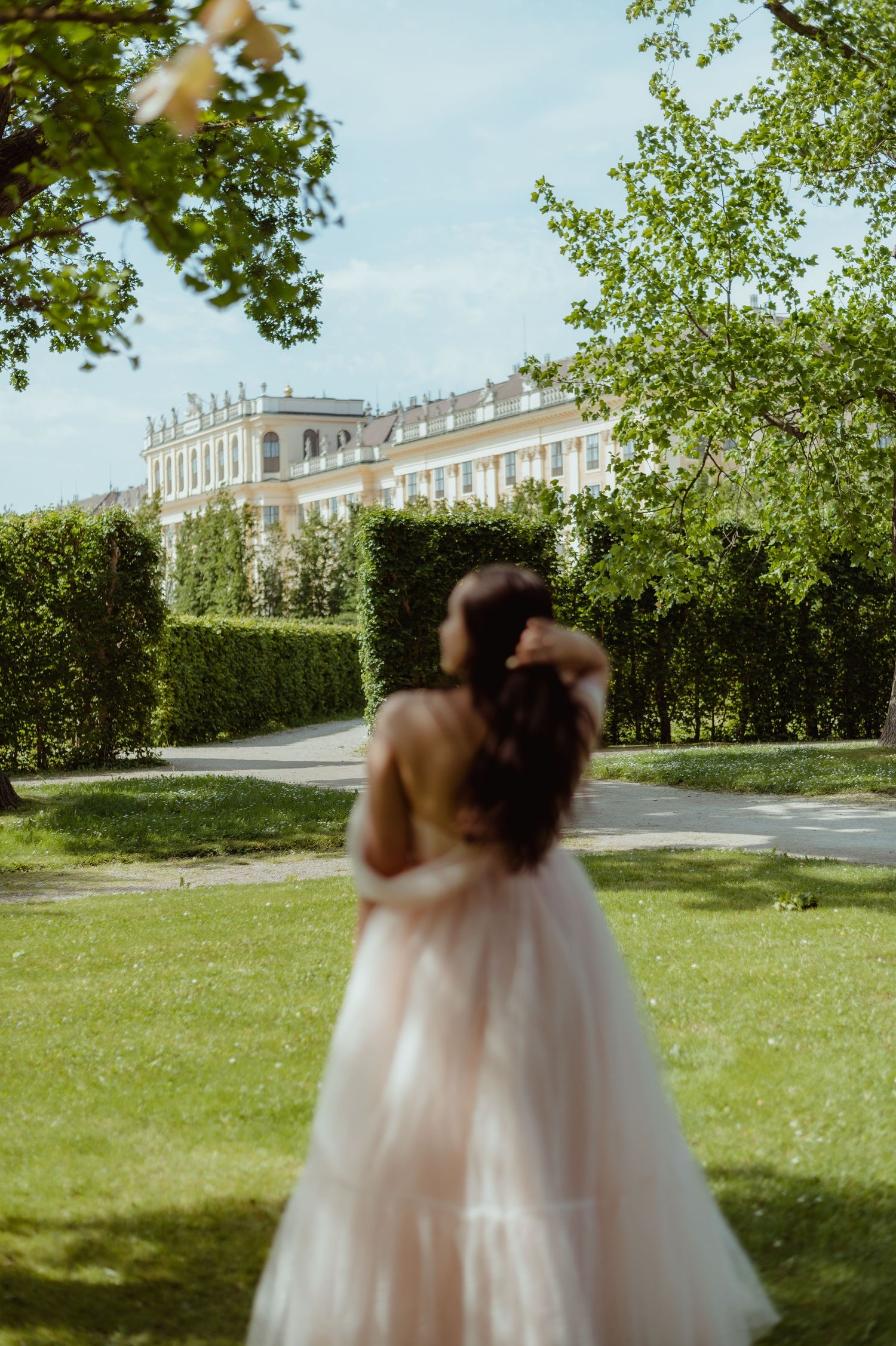 Woman in blush pink tulle gown stands in manicured garden with grand palace building and green hedges in background.