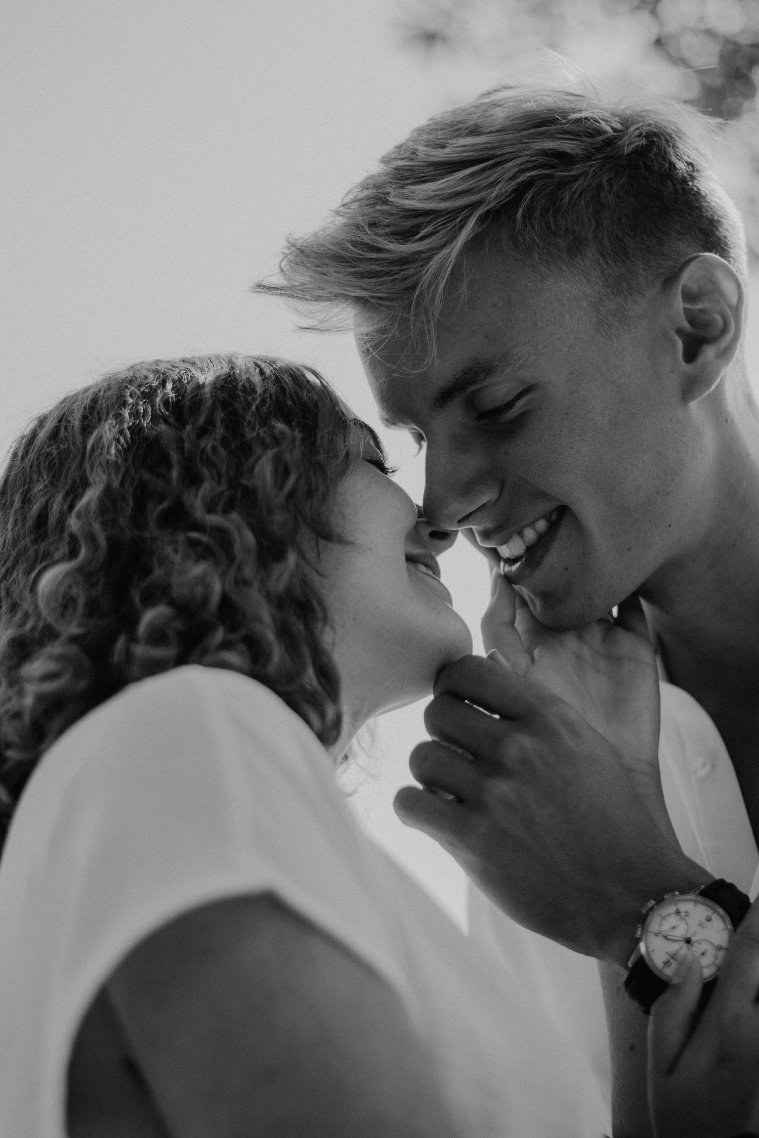 Black and white photo of a smiling couple sharing a tender moment, almost kissing, man wearing a watch.