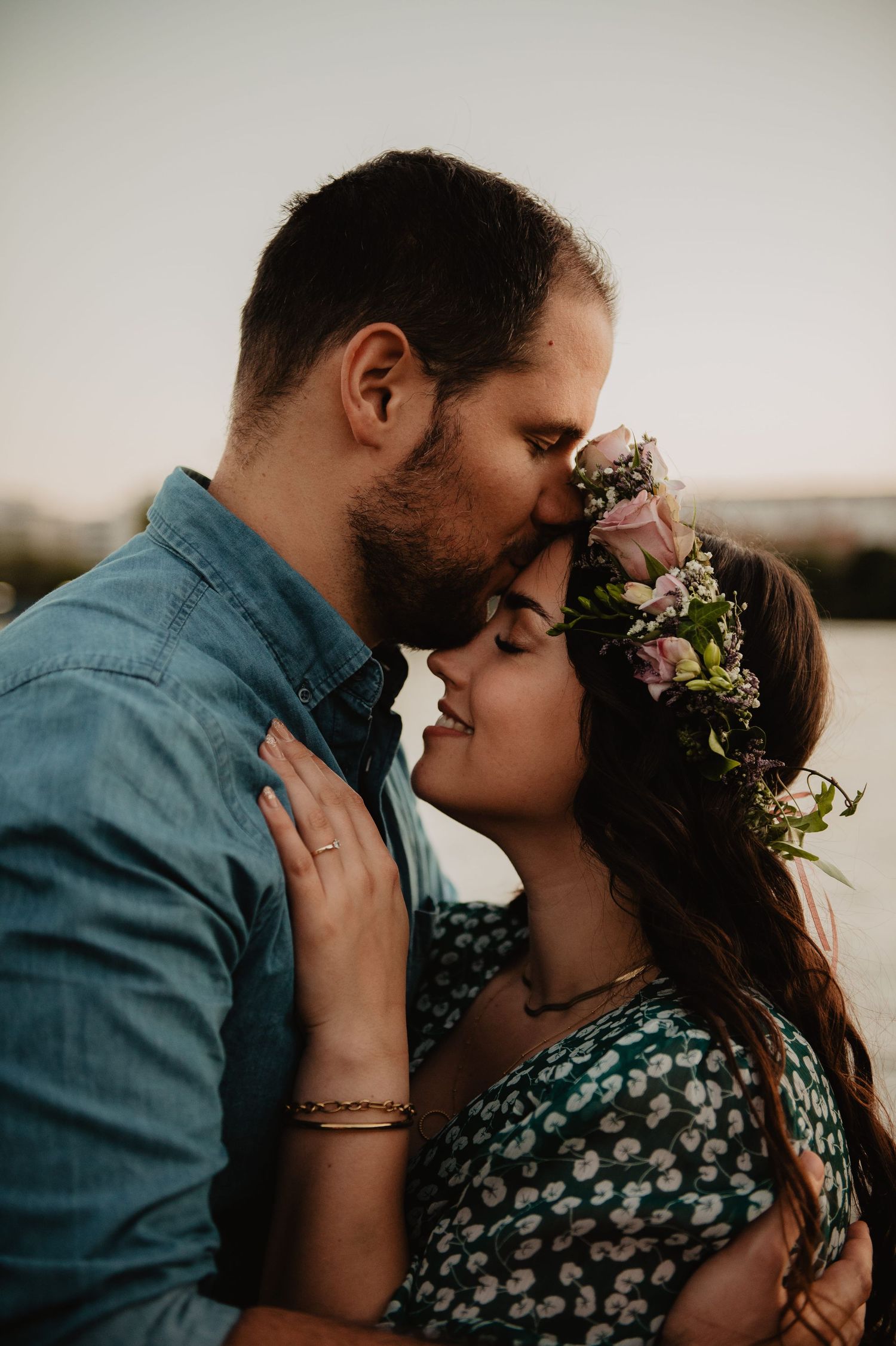 Couple sharing a tender forehead kiss at sunset, woman wearing a floral crown with pink roses.