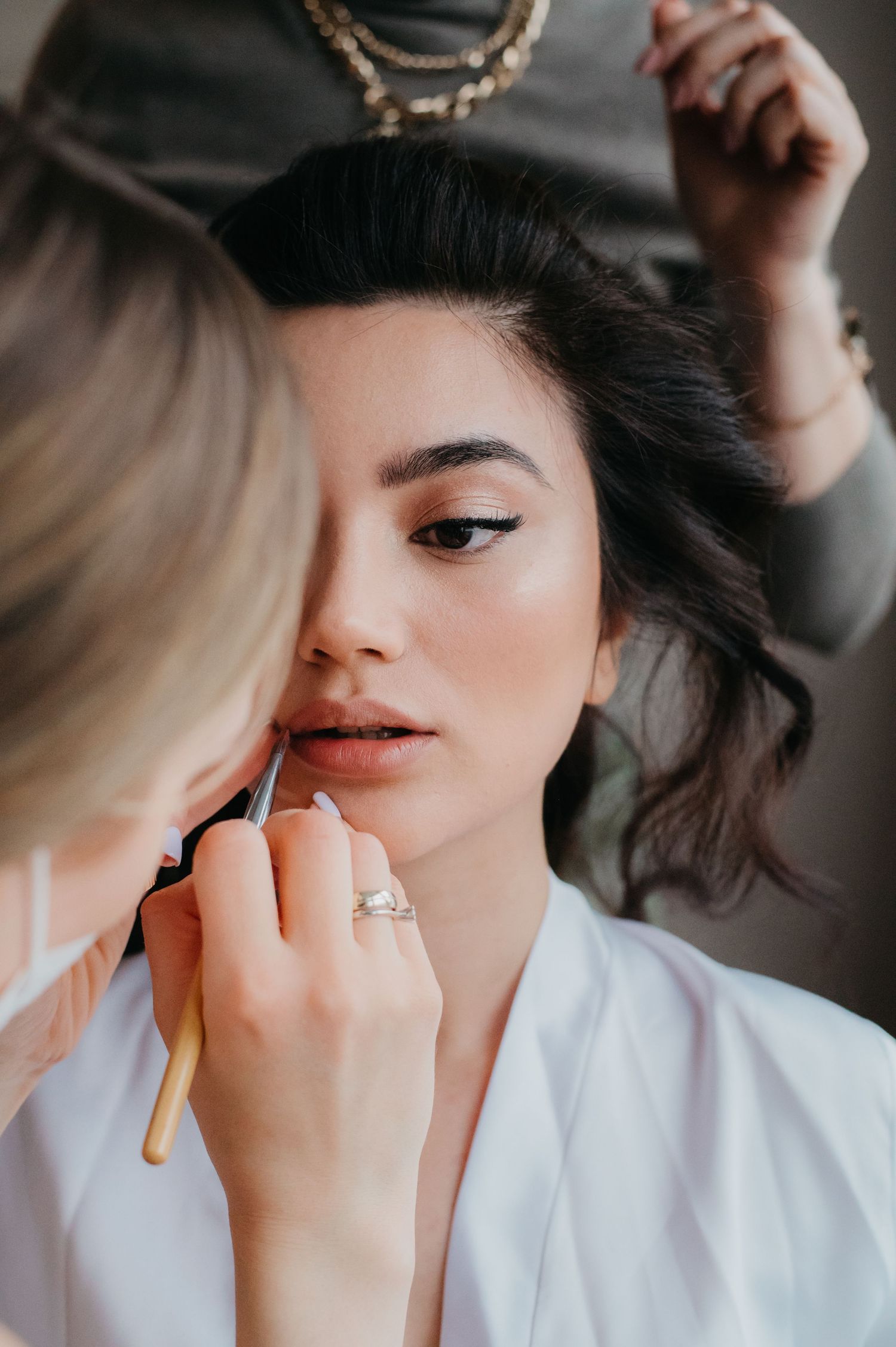 A makeup artist applies lip color to a dark-haired bride in a white robe during wedding preparations.