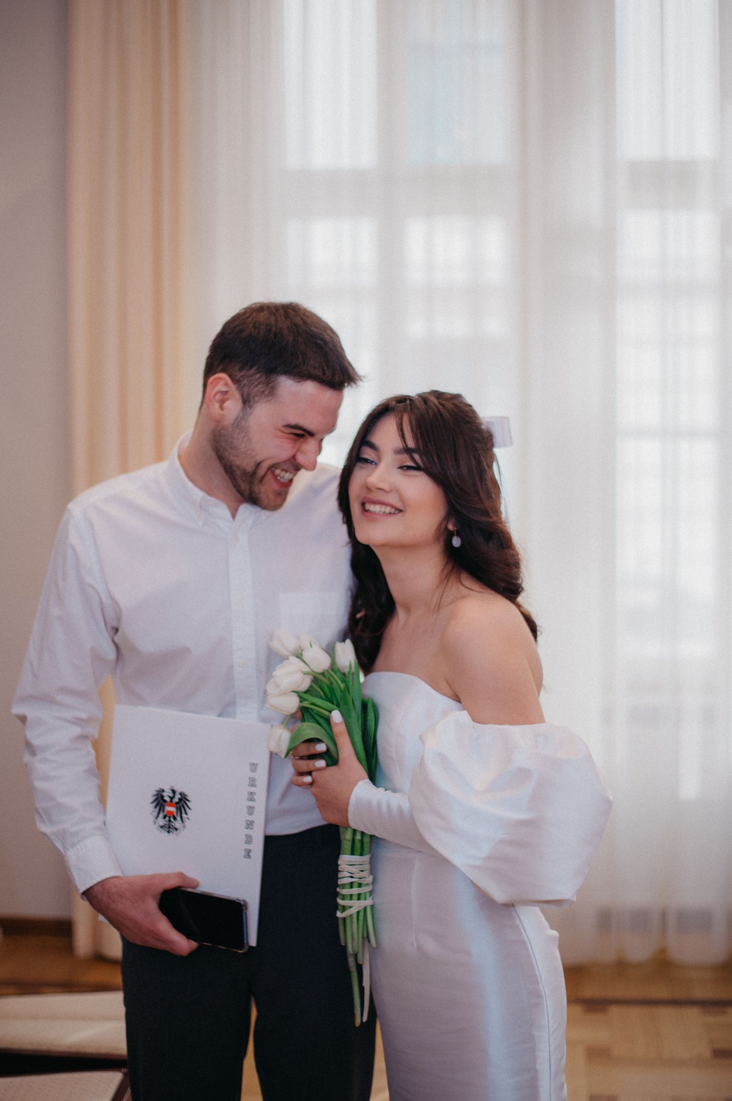Newlywed couple in white attire smiling, holding tulips and marriage certificate at civil wedding ceremony.