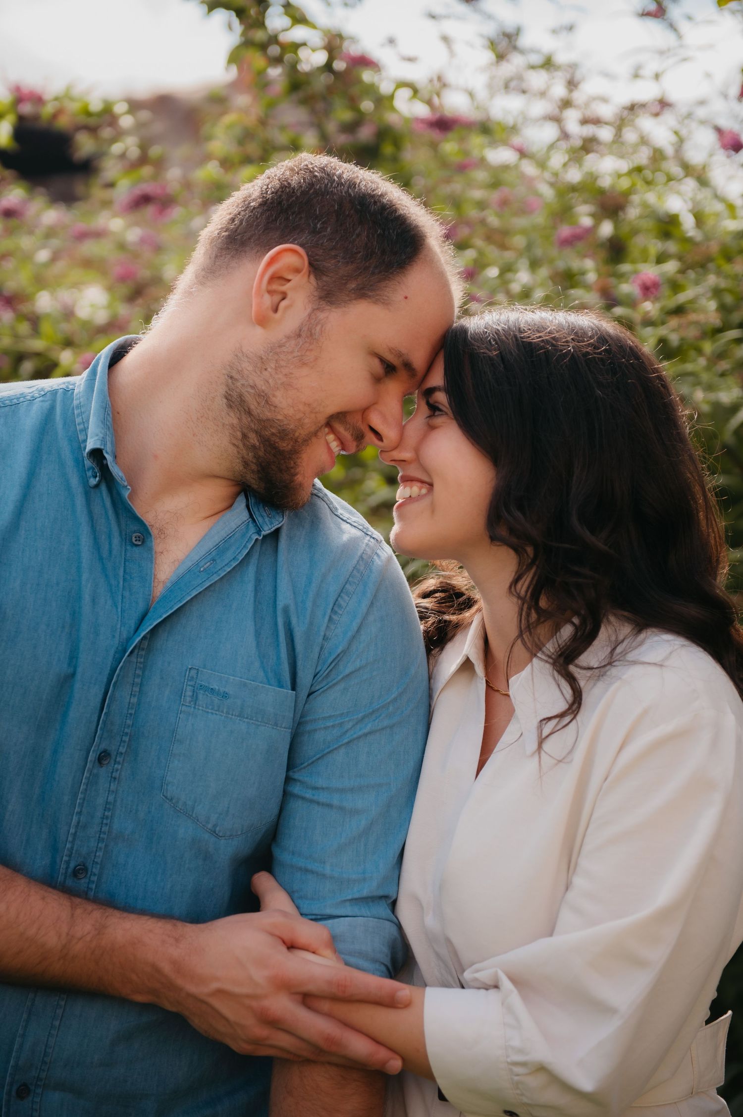 A couple smiles and touches foreheads in a romantic outdoor embrace surrounded by blooming flowers.