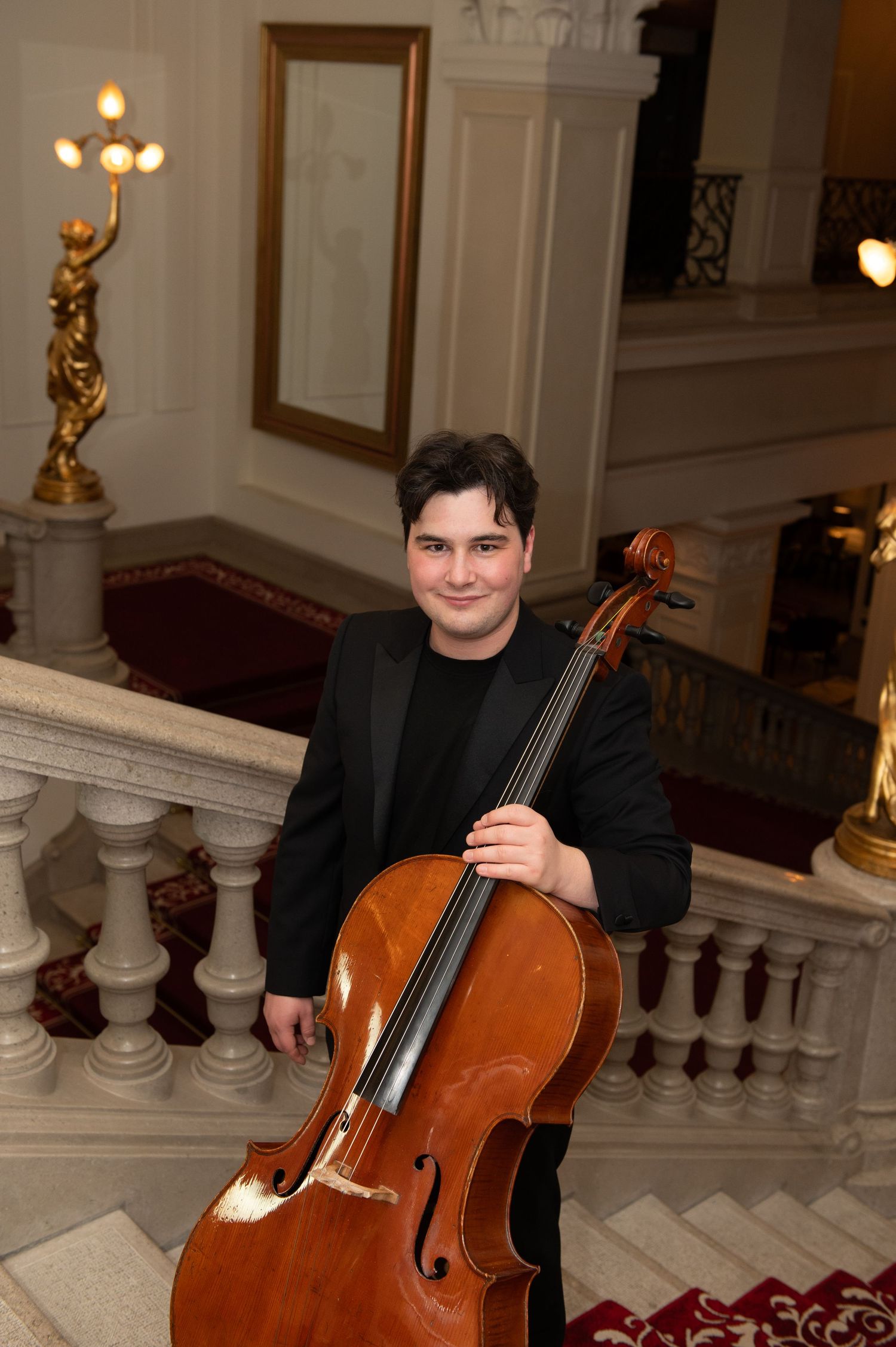 Young male cellist in black jacket smiling while holding cello on elegant staircase with gold candelabra.