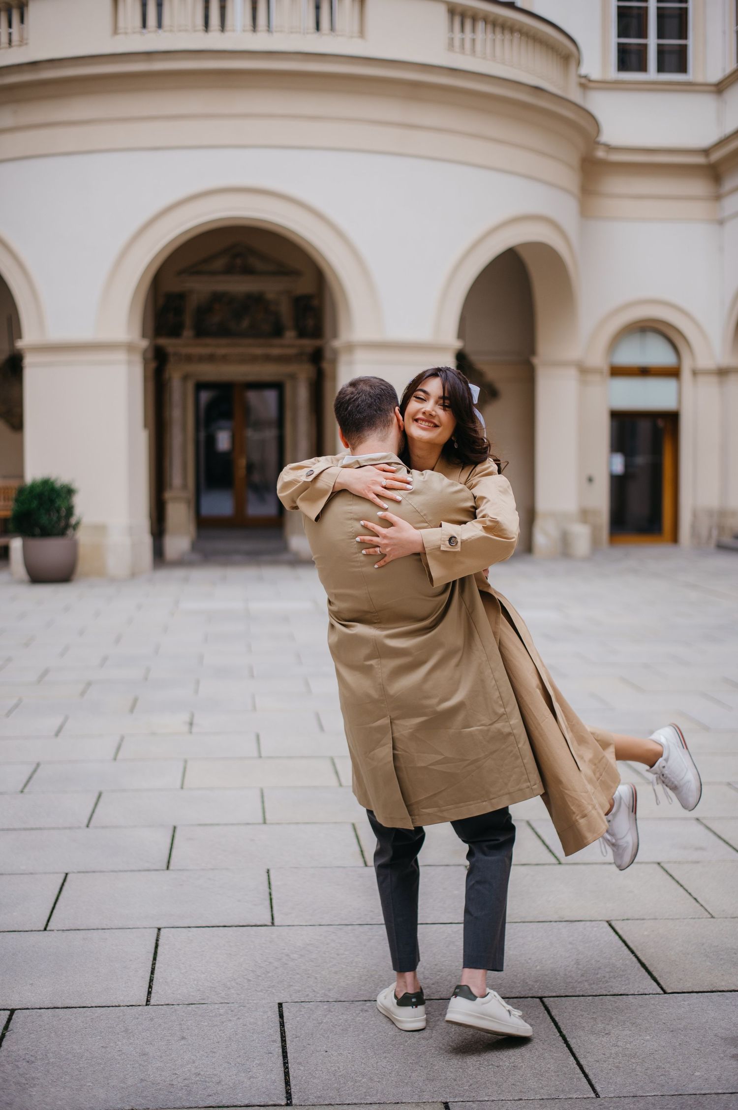Joyful couple embracing and spinning in front of a grand neoclassical building on a cobblestone plaza.