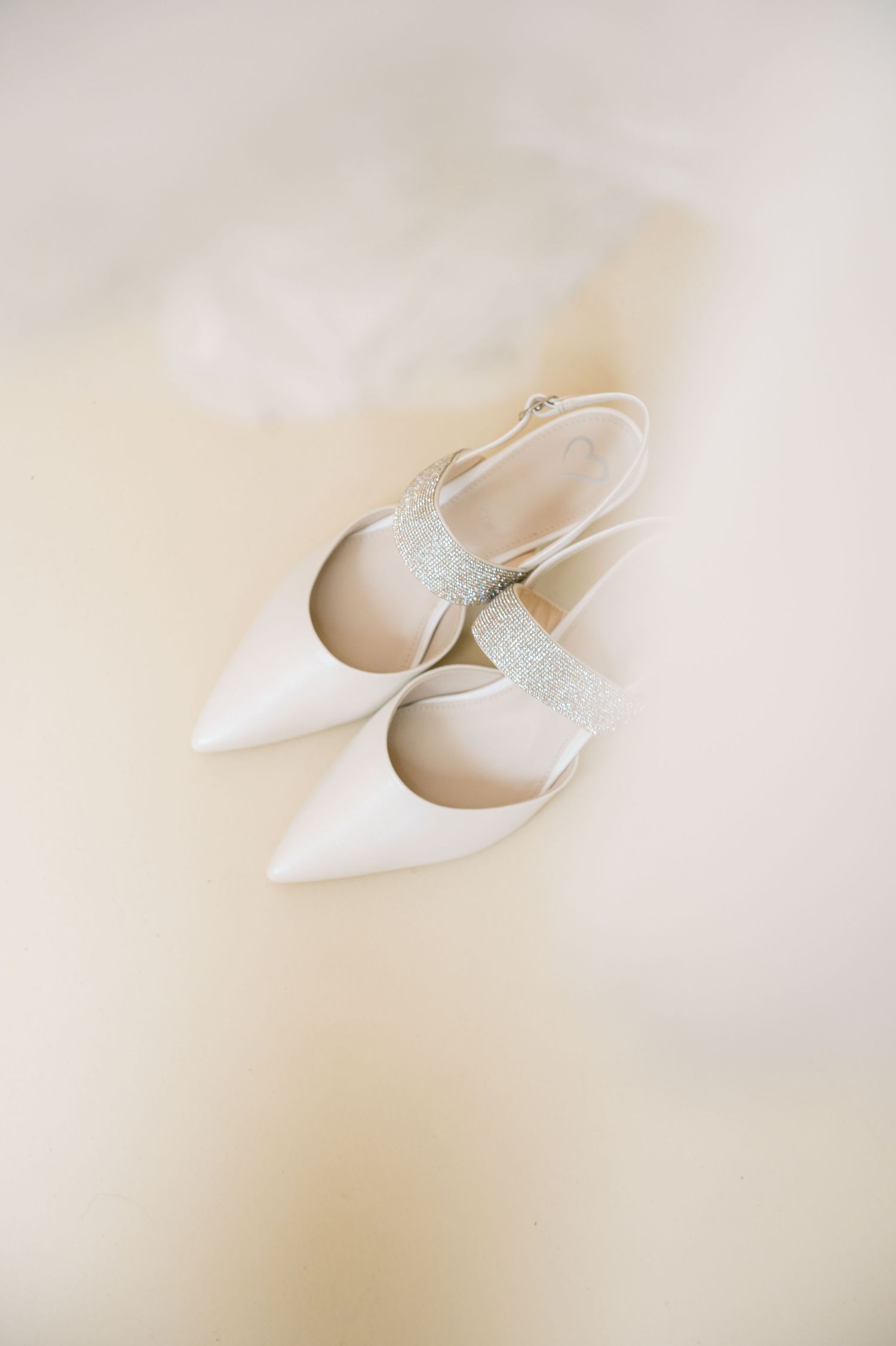 Elegant white satin pointed-toe bridal heels with sparkling glitter ankle strap on a soft ivory background.