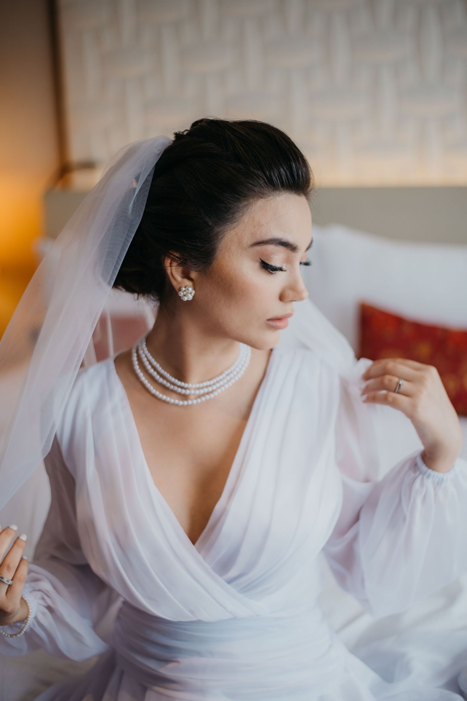 Elegant bride in white chiffon gown with pearl necklace and earrings, wearing a veil, seated and looking down gracefully.
