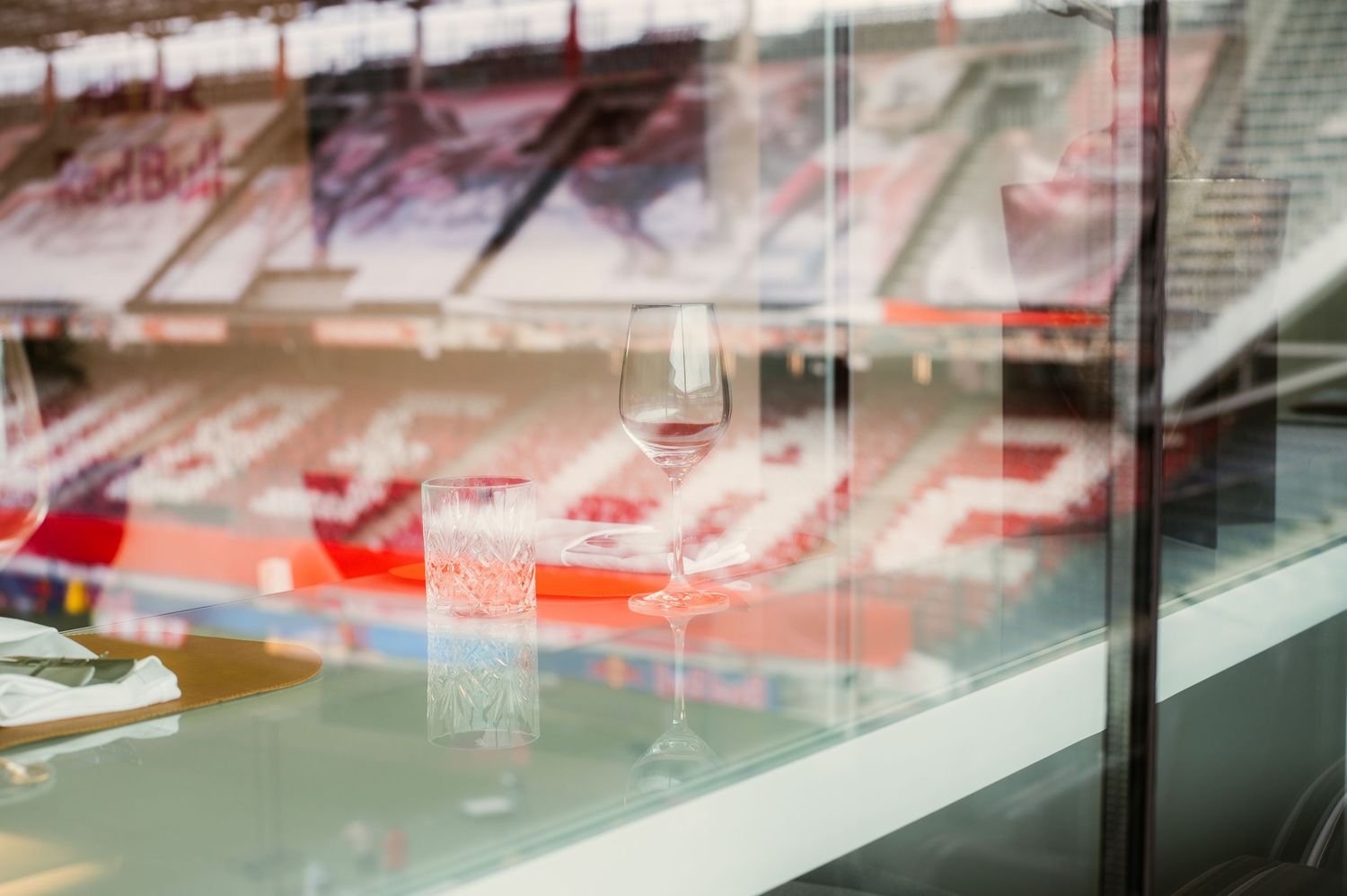 Wine glass on a glass ledge overlooking empty red stadium seats in a modern sports arena.