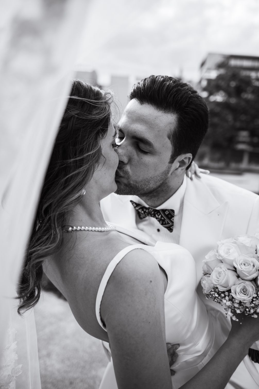 Black and white photo of bride and groom sharing a romantic kiss under a flowing veil outdoors.