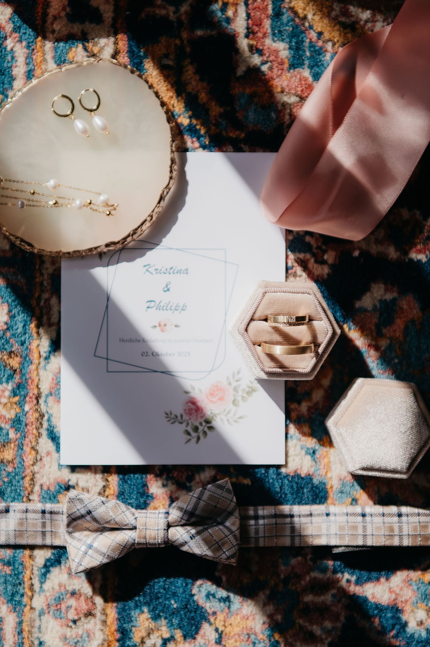 Wedding flat lay with invitation, rings in velvet box, bow tie, earrings, and gold jewelry on a colorful rug.