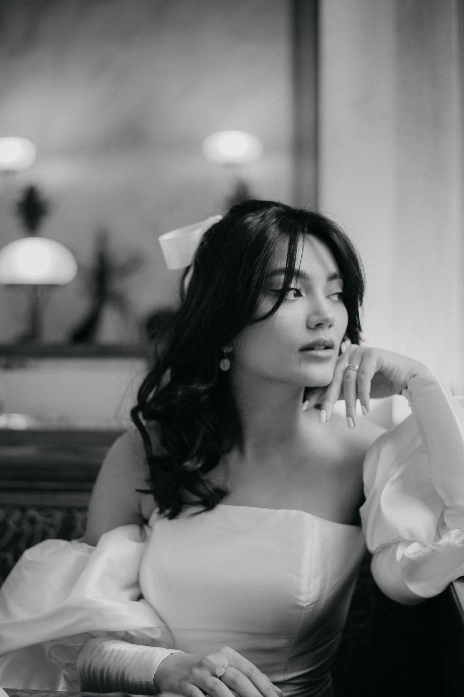 Black and white portrait of an elegant woman in a white off-shoulder gown with a bow, posed thoughtfully in a luxurious interior.