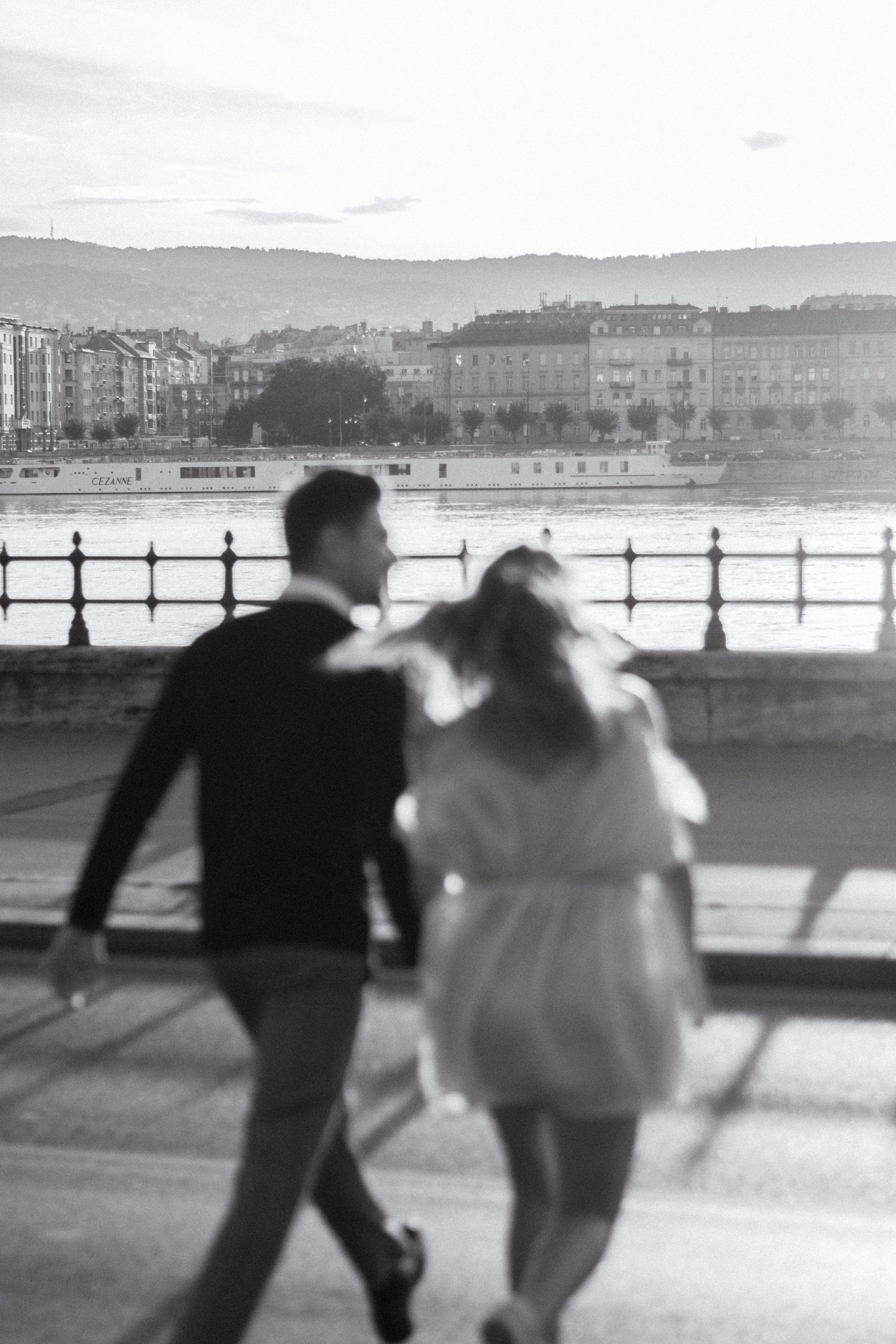 Black and white photo of a couple walking along a riverside promenade with city buildings in the background.