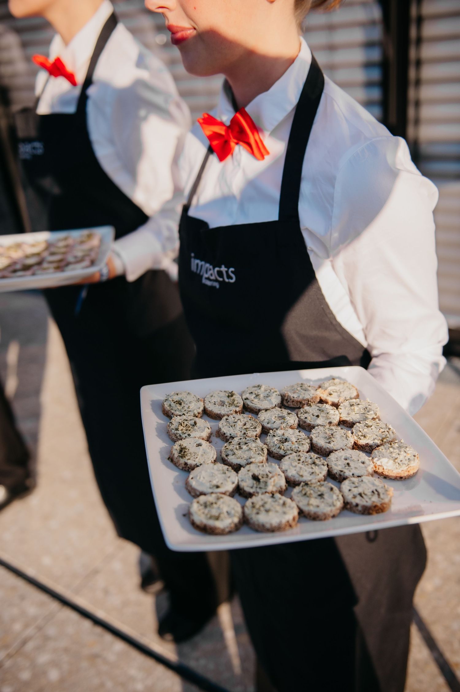 Catering staff in black Impacts aprons and red bow ties serving trays of bite-sized appetizers at an event.