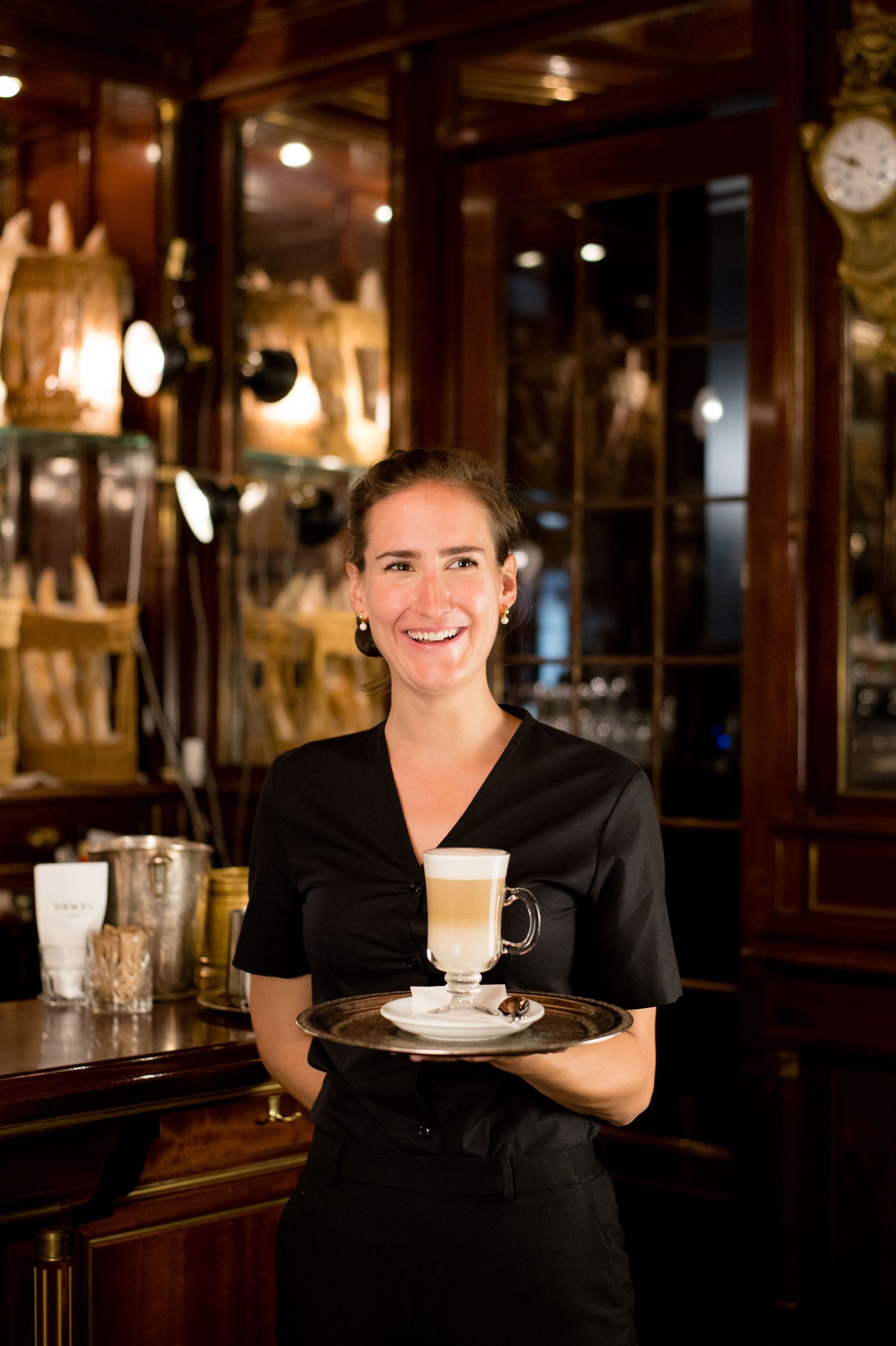 Smiling waitress in black uniform holding a silver tray with a latte in an elegant restaurant.